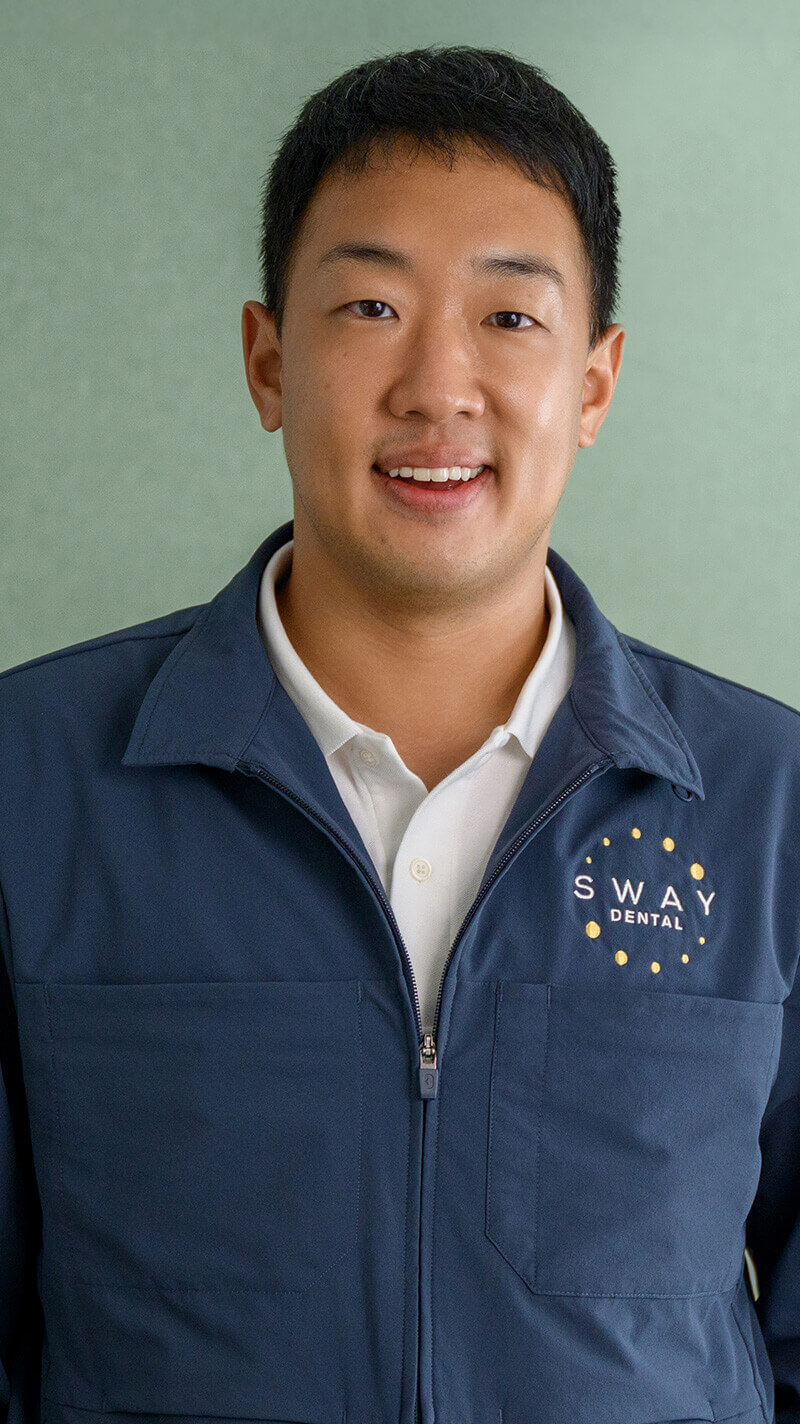 Smiling young man wearing a navy blue jacket with Sway Dental logo over a white collared shirt against a light green background.