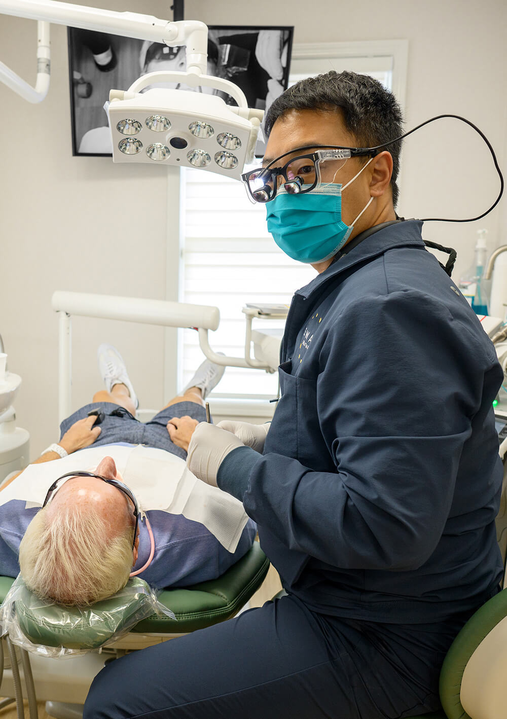 Dentist wearing magnifying glasses and a teal mask treating an elderly male patient reclining in a dental chair.