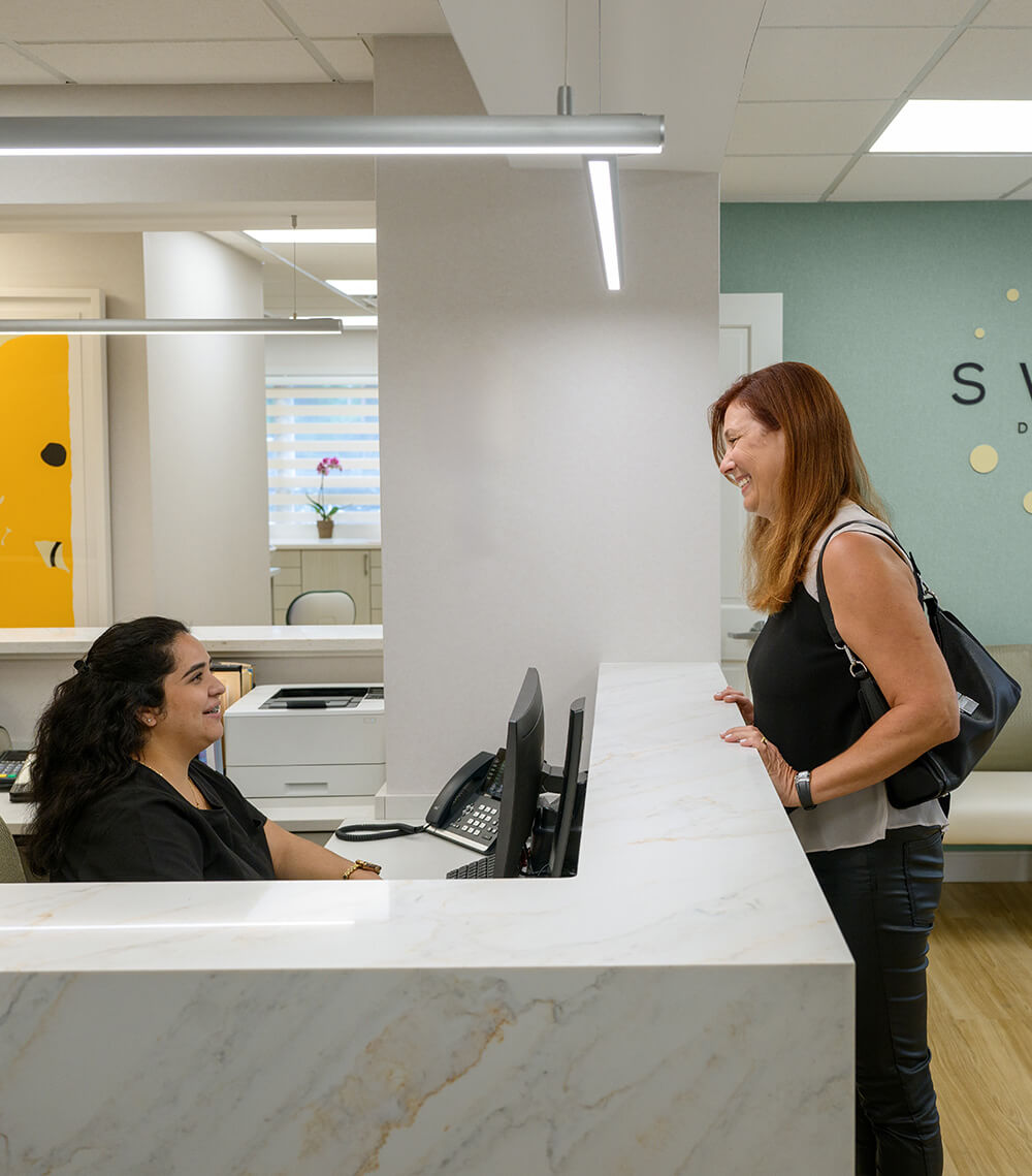 Smiling receptionist talking to a standing woman at a marble reception desk in a modern office.