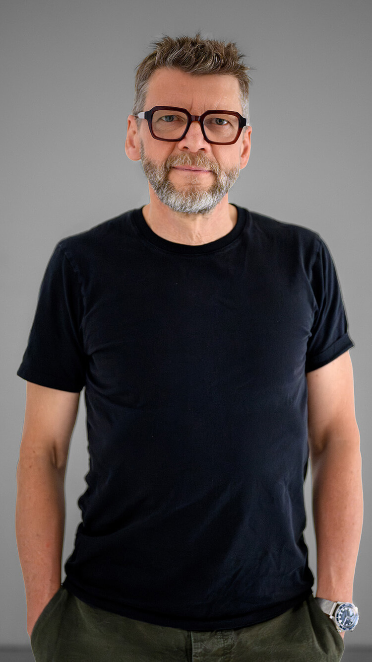 CNR Group luxury builders team member: Jesse. Middle-aged man with short graying hair, beard, and large glasses wearing a black t-shirt and watch, standing against a gray background.