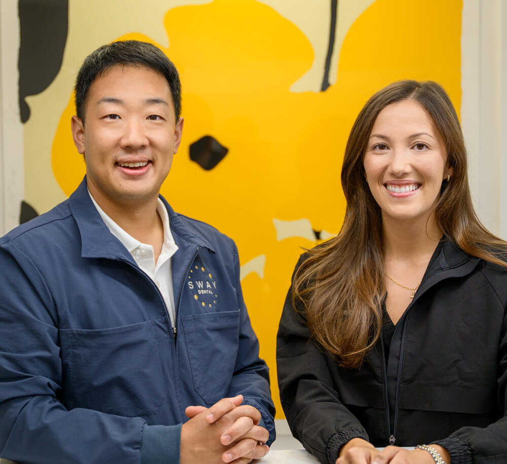 Smiling man and woman sitting side by side in front of a yellow and black abstract wall art.