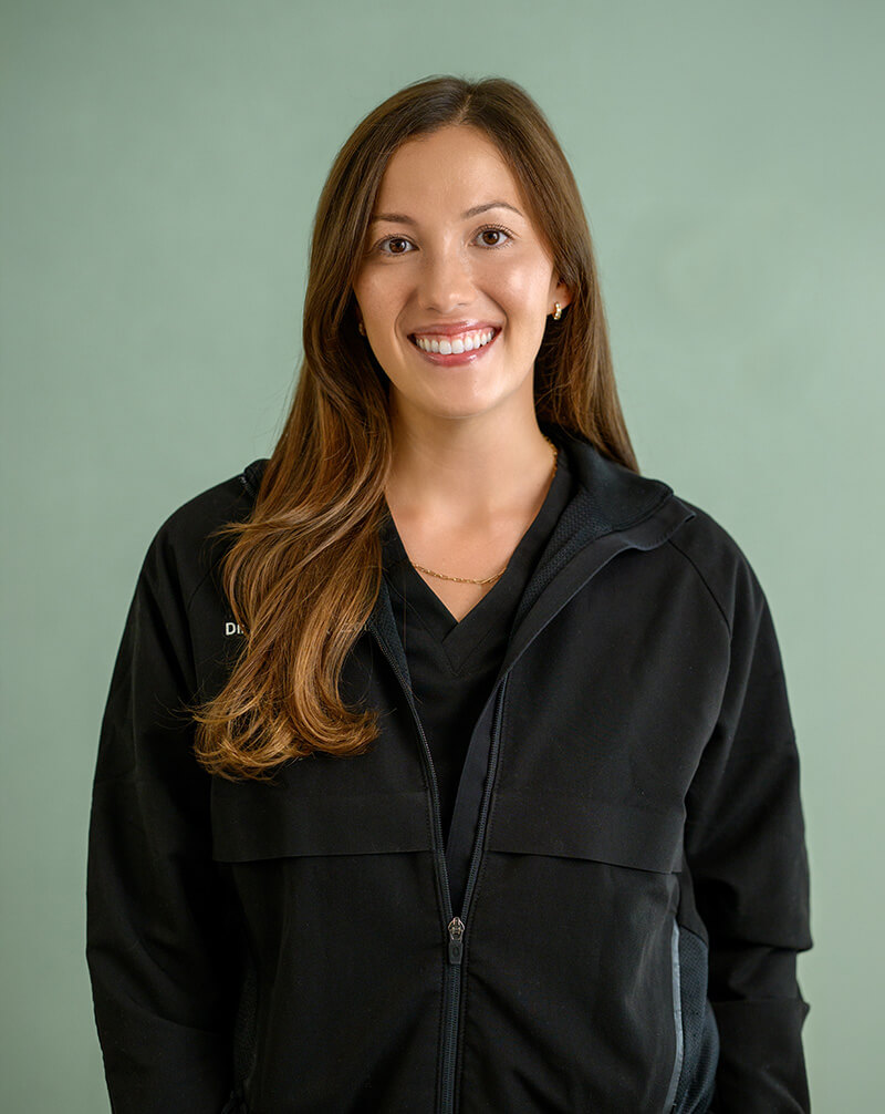 Smiling woman with long brown hair wearing a black zip-up jacket over a black top, standing against a green background.