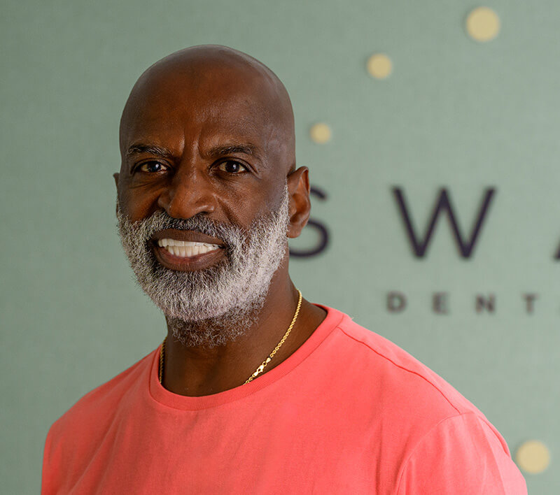 Smiling bald man with a gray beard wearing a coral shirt and gold necklace in front of a green wall with partial text.
