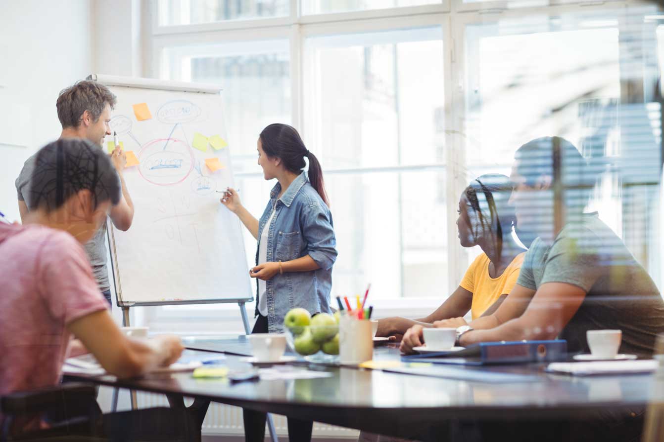 Four people in a bright office, one woman is presenting and writing on a flip chart while others listen attentively.