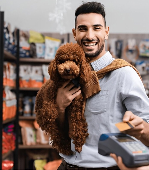 Smiling man holding a brown poodle and presenting a credit card at a payment terminal in a pet store.