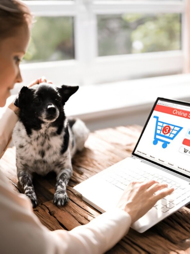 Person using a white laptop with an online shopping cart displayed, sitting next to a black and white dog on a wooden table by a window.