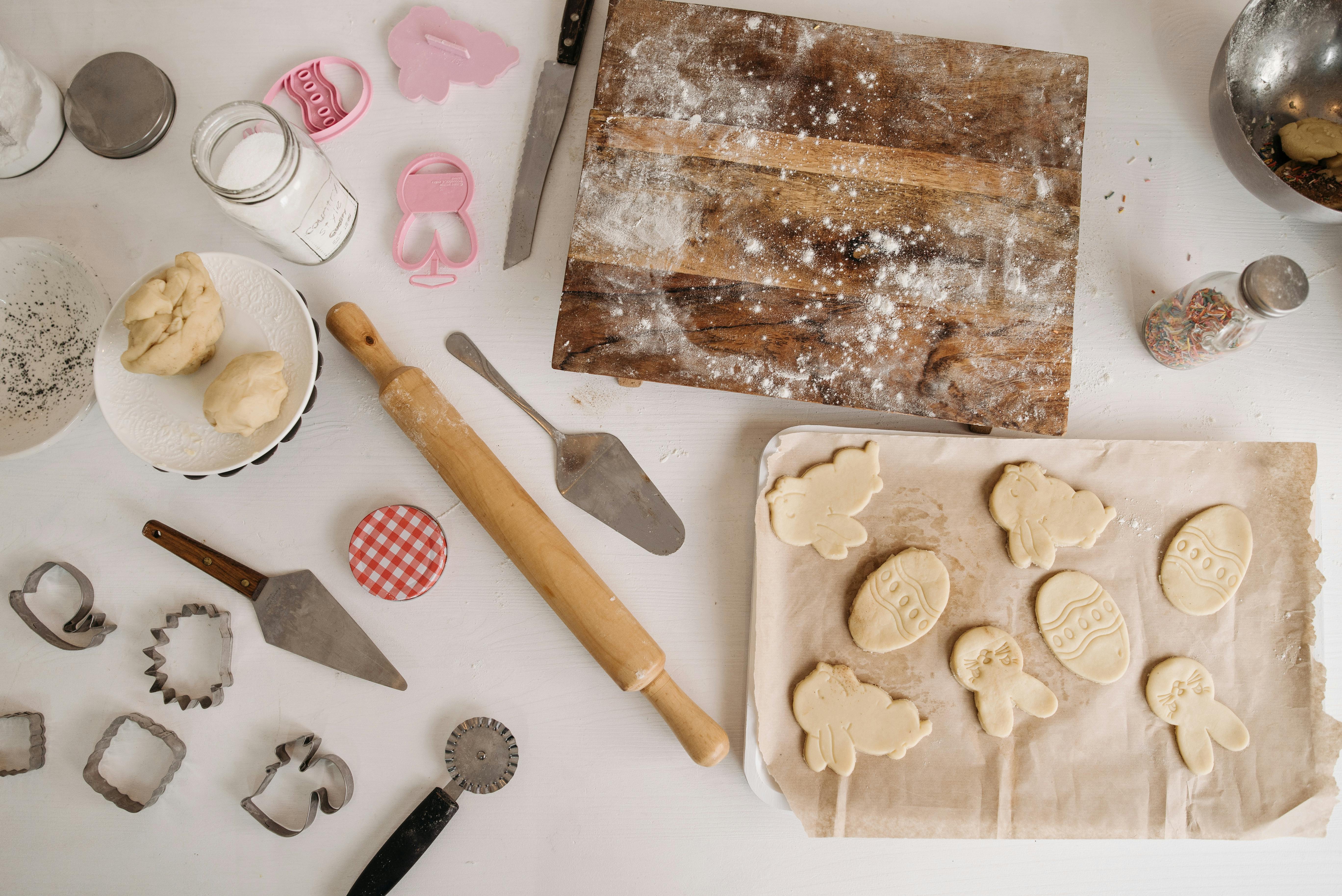 Baking setup with wooden rolling pin, cookie cutters, dough on a white plate, and cut-out Easter-themed cookies on parchment paper.