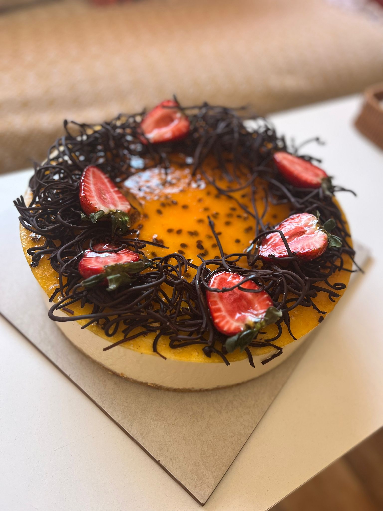 Round cheesecake topped with passion fruit glaze, chocolate shavings, and halved strawberries on a cardboard surface.