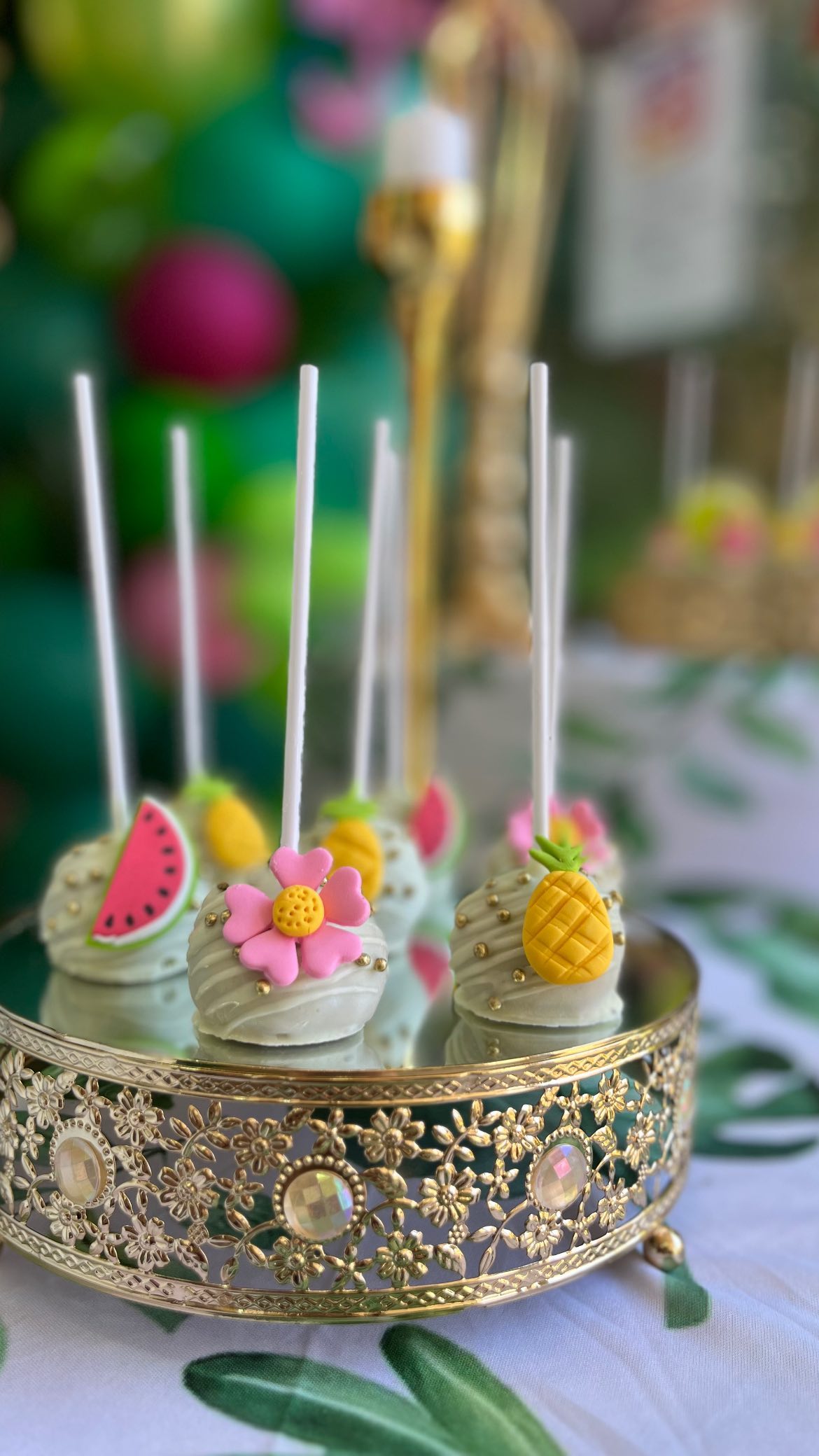 Decorative cake pops with white coating and tropical-themed fondant toppers including pineapples, watermelon slices, and pink flowers on an ornate gold cake stand.