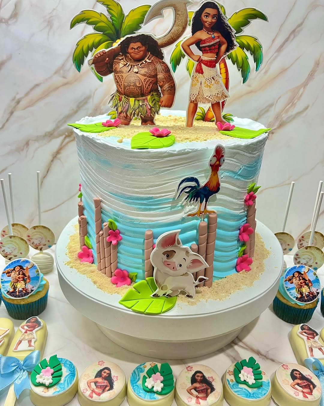 Moana-themed cake with characters Maui and Moana figurines, decorated with palm trees, Pua the pig, Hei Hei the rooster, pink flowers, and matching themed cupcakes and treats.