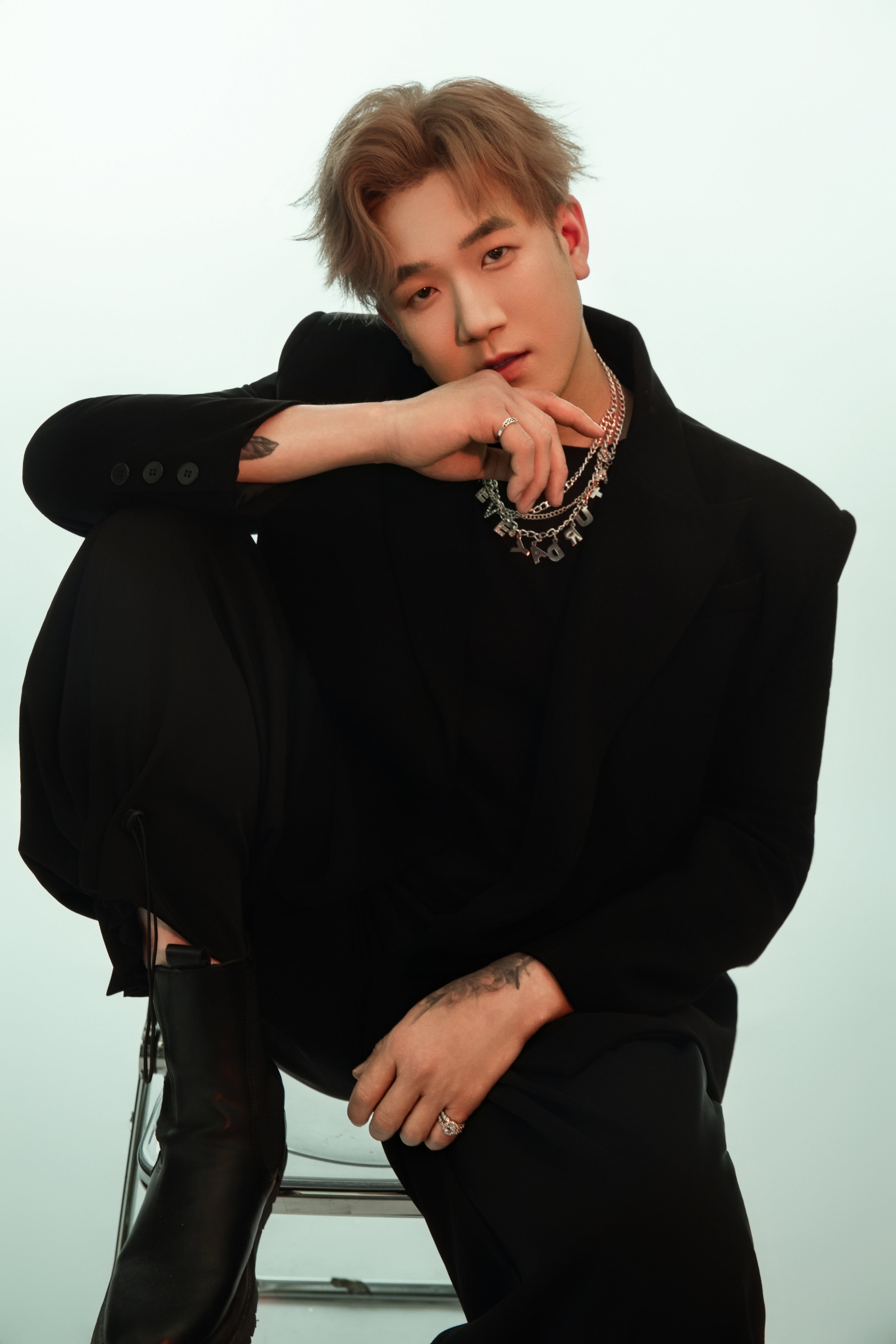 Seated on a chair with one leg bent atop it, a man gazes at the camera.