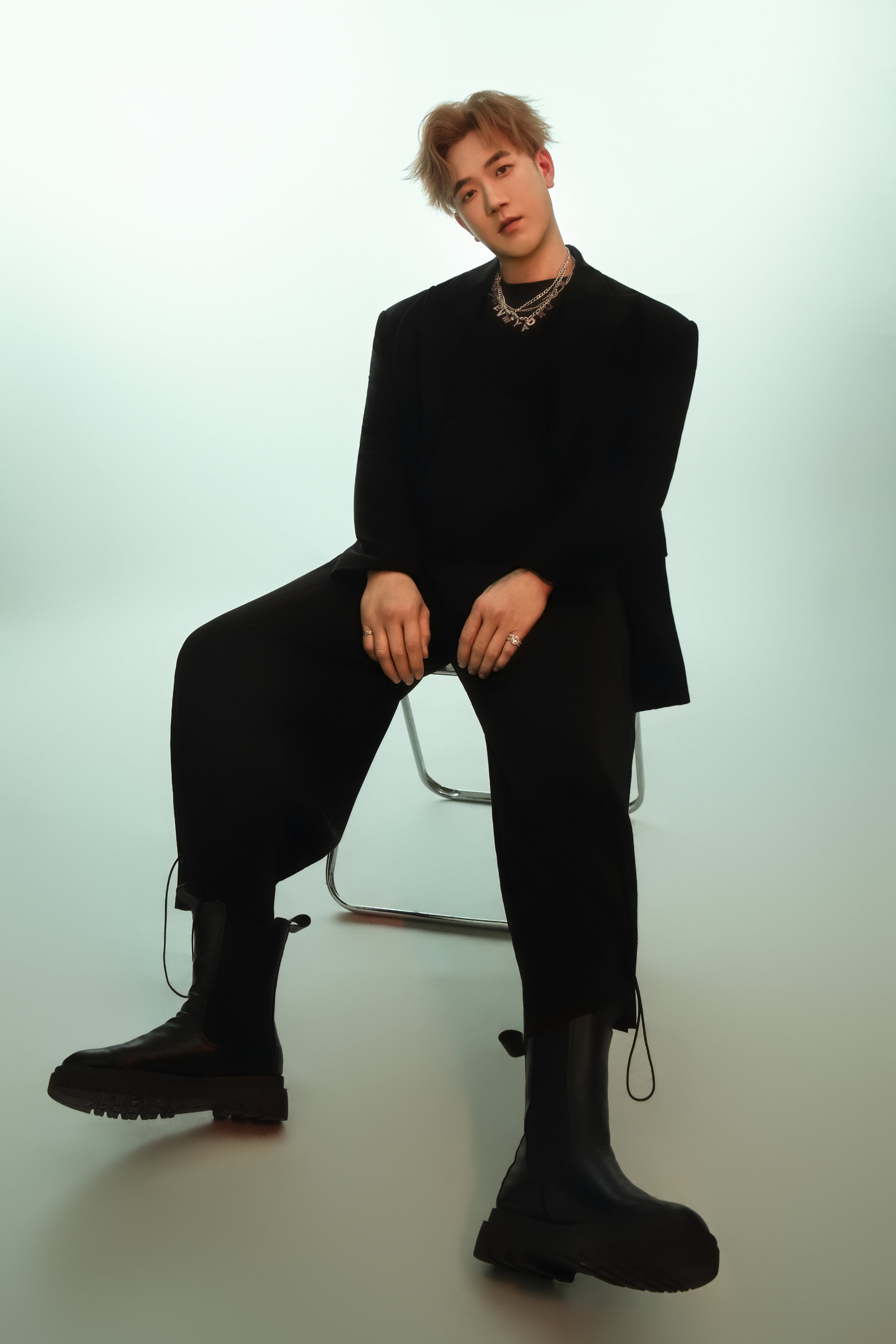 A man sits on a chair in a relaxed pose, legs stretched out, with an emerald-green tinted background.