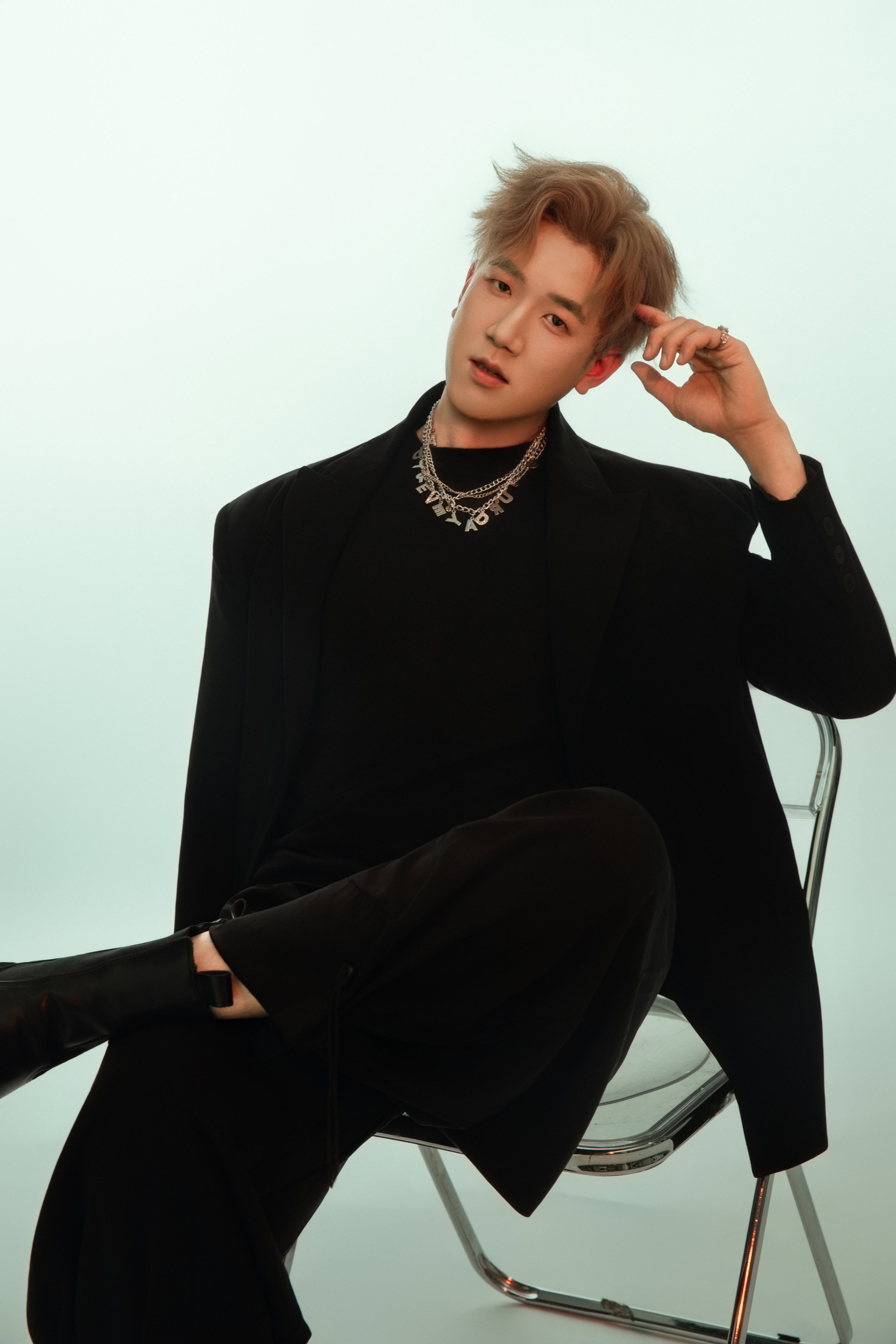 Seated on a chair, a man gazes at the camera, his arm propped on the chair holding up his head.