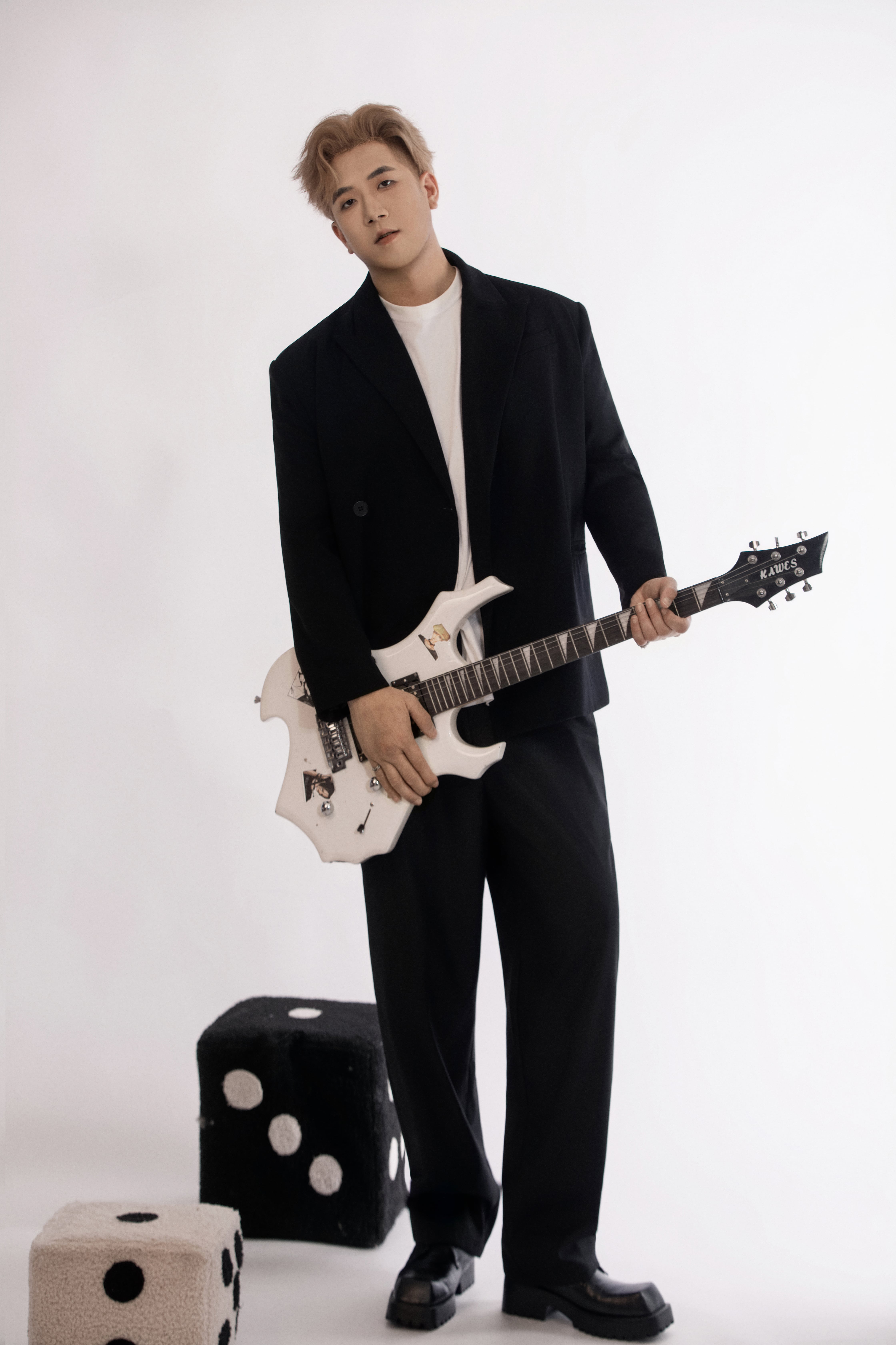 A man in a black suit holds a white electric guitar in a minimalist studio, standing near oversized dice against a white background.
