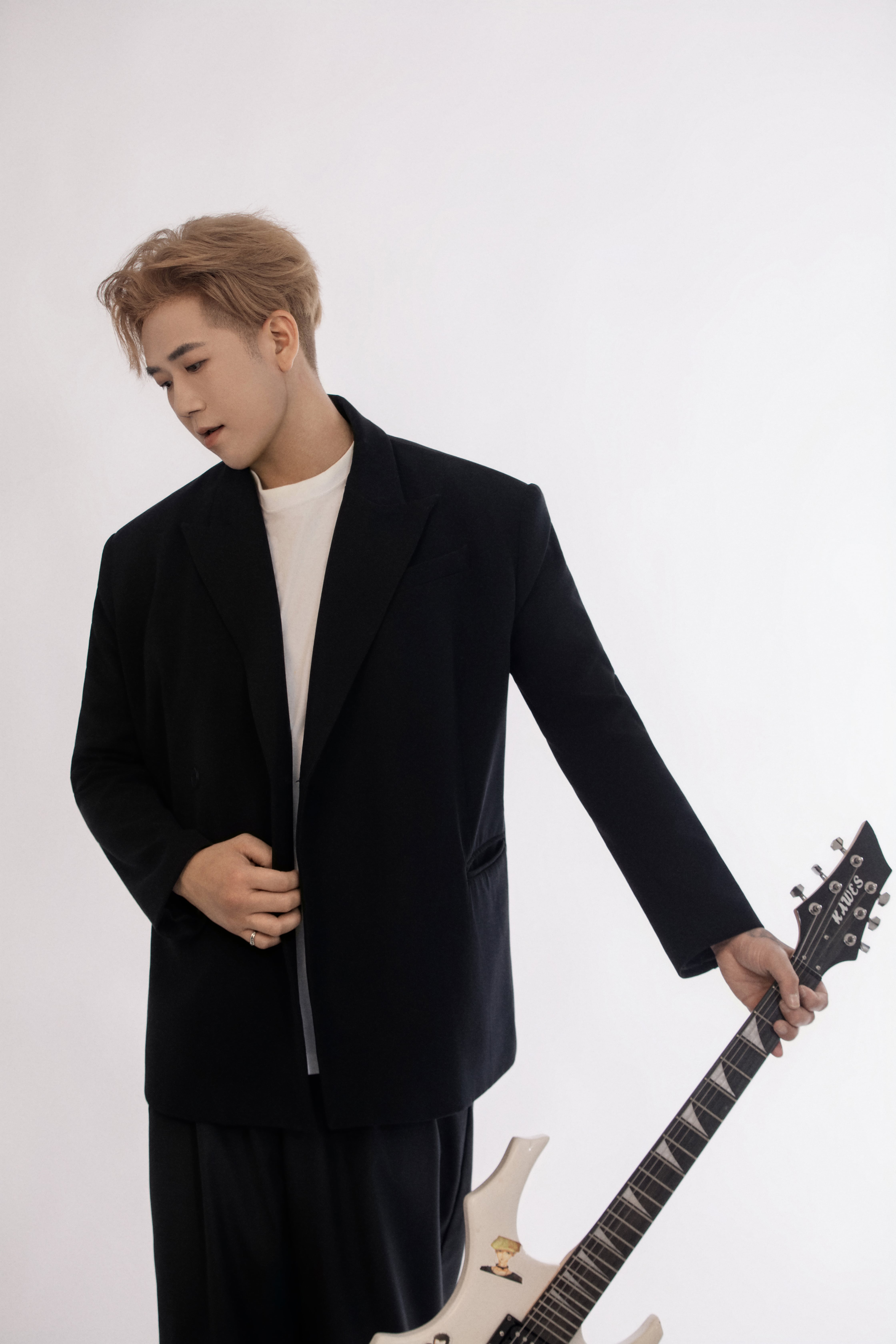 A man wearing a black suit holds a white electric guitar in a studio setting, looking away from the camera.