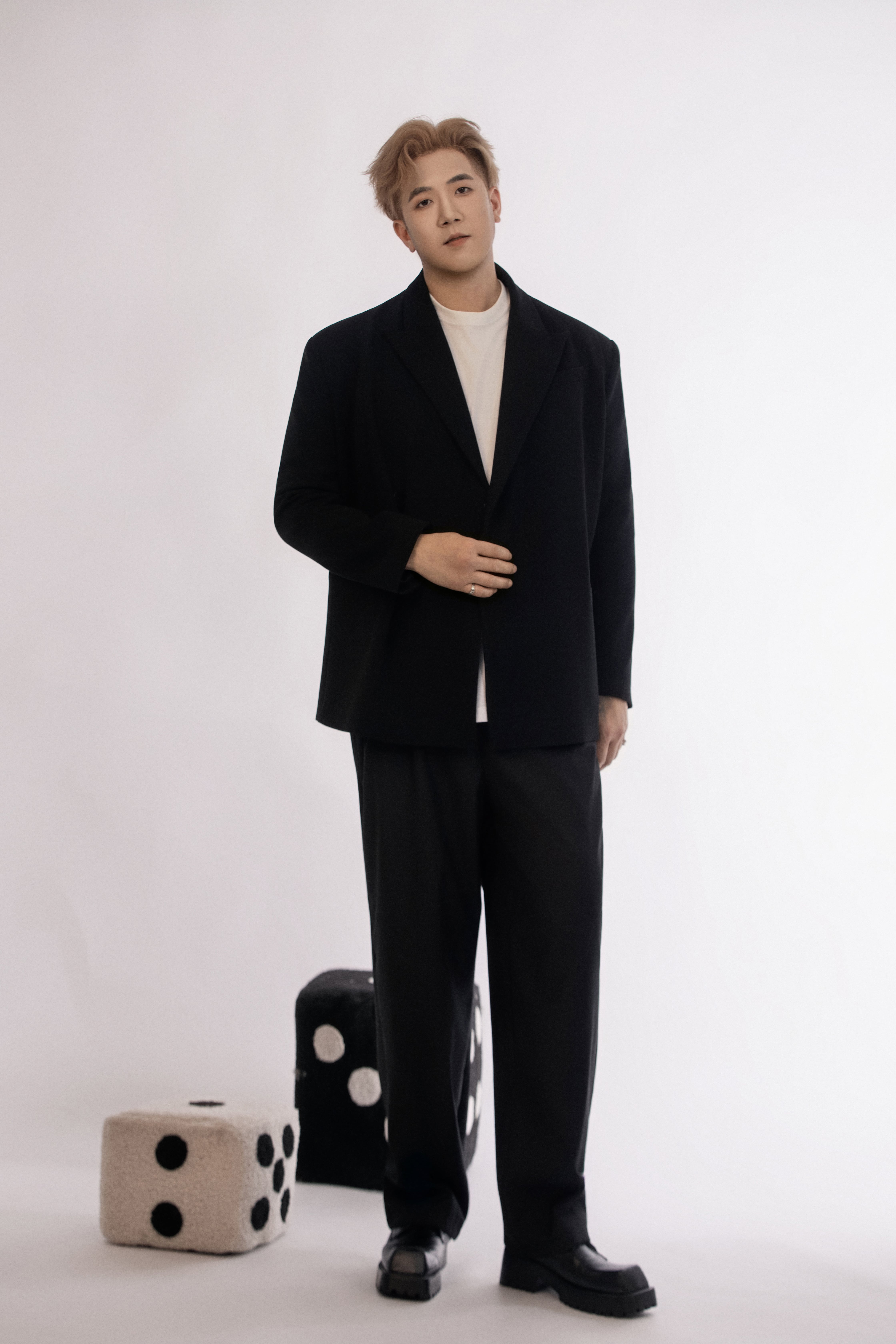 A man stands in a minimalist studio wearing a black suit and white shirt, posed casually beside oversized black and white dice.