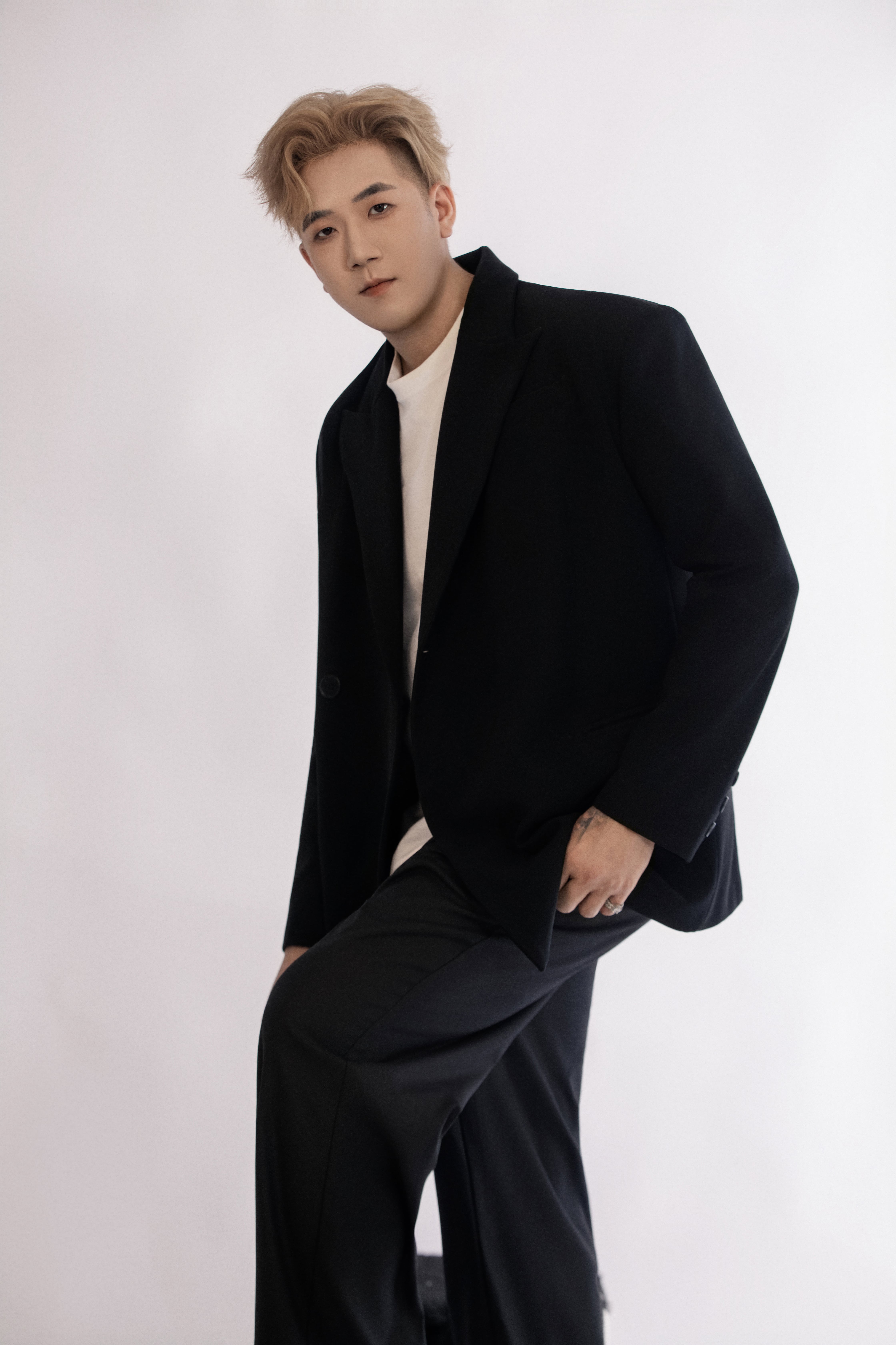 A man in a black suit leans slightly forward in a white studio space, looking directly at the camera with a relaxed expression.