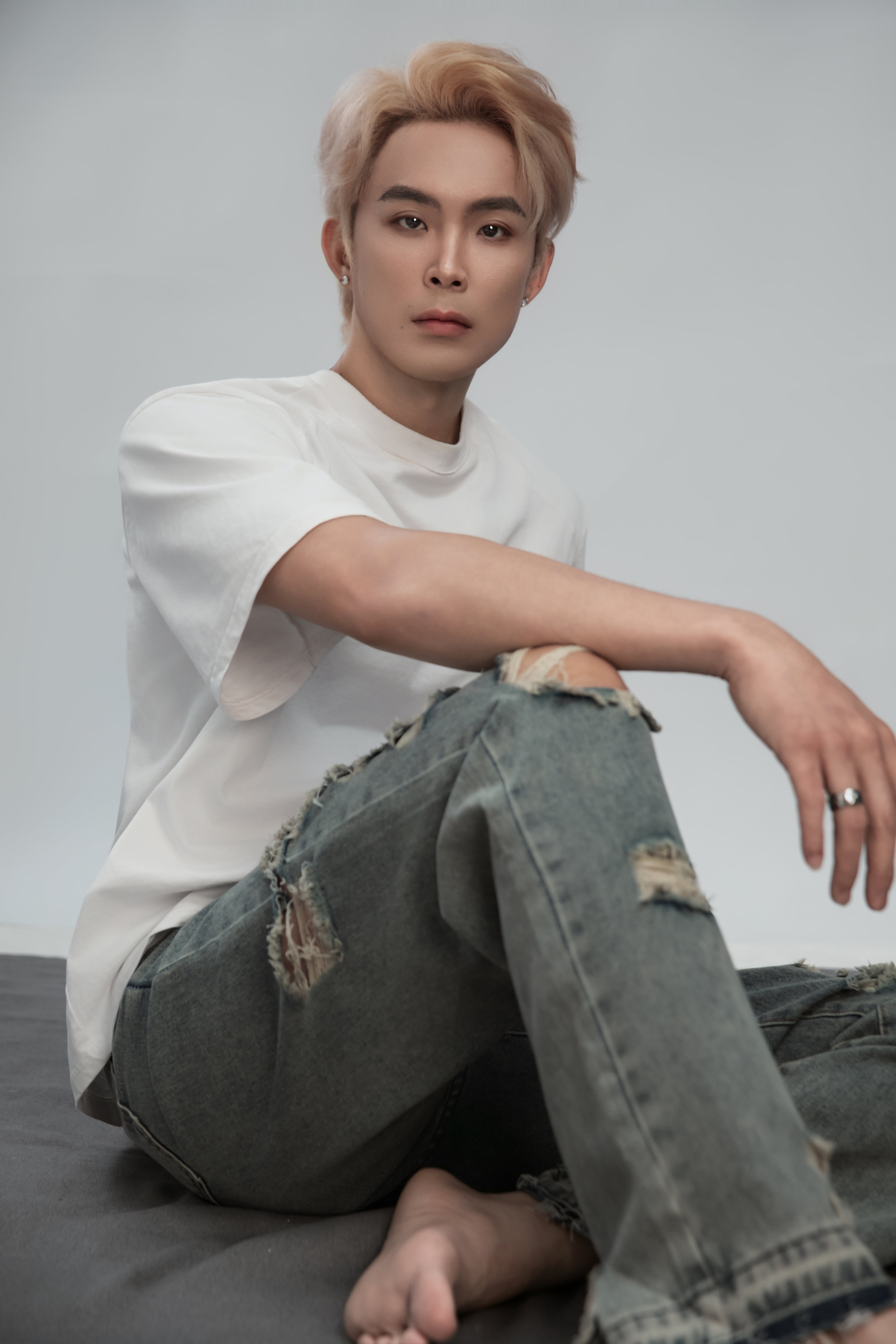 A man in a white shirt and jeans sits with his legs extended, one crossed over the other, gazing directly at the camera against a white backdrop with a gray rug beneath him.