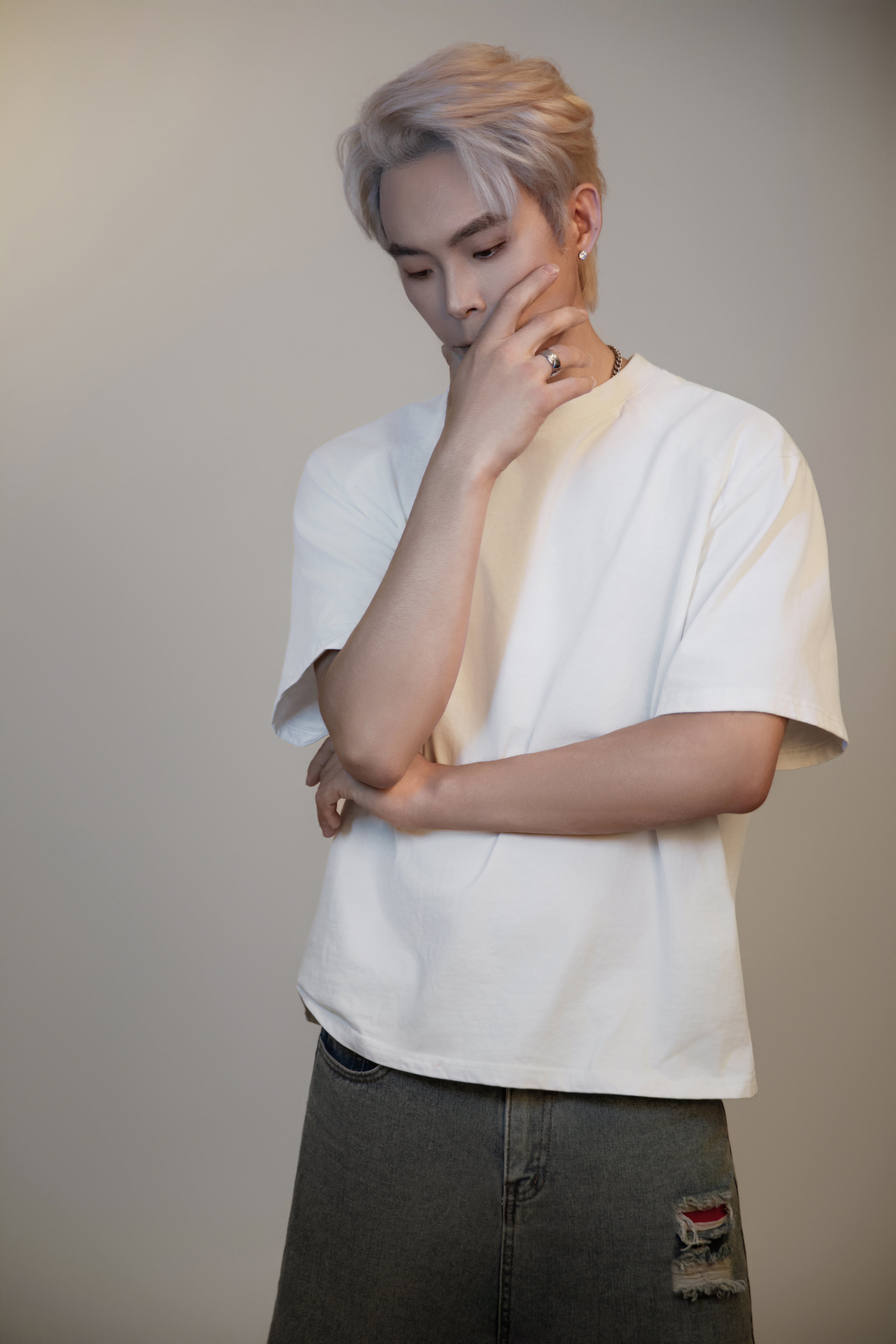 A man poses in front of a gray background, one arm resting across his face and the other holding it, with his gaze directed downward.