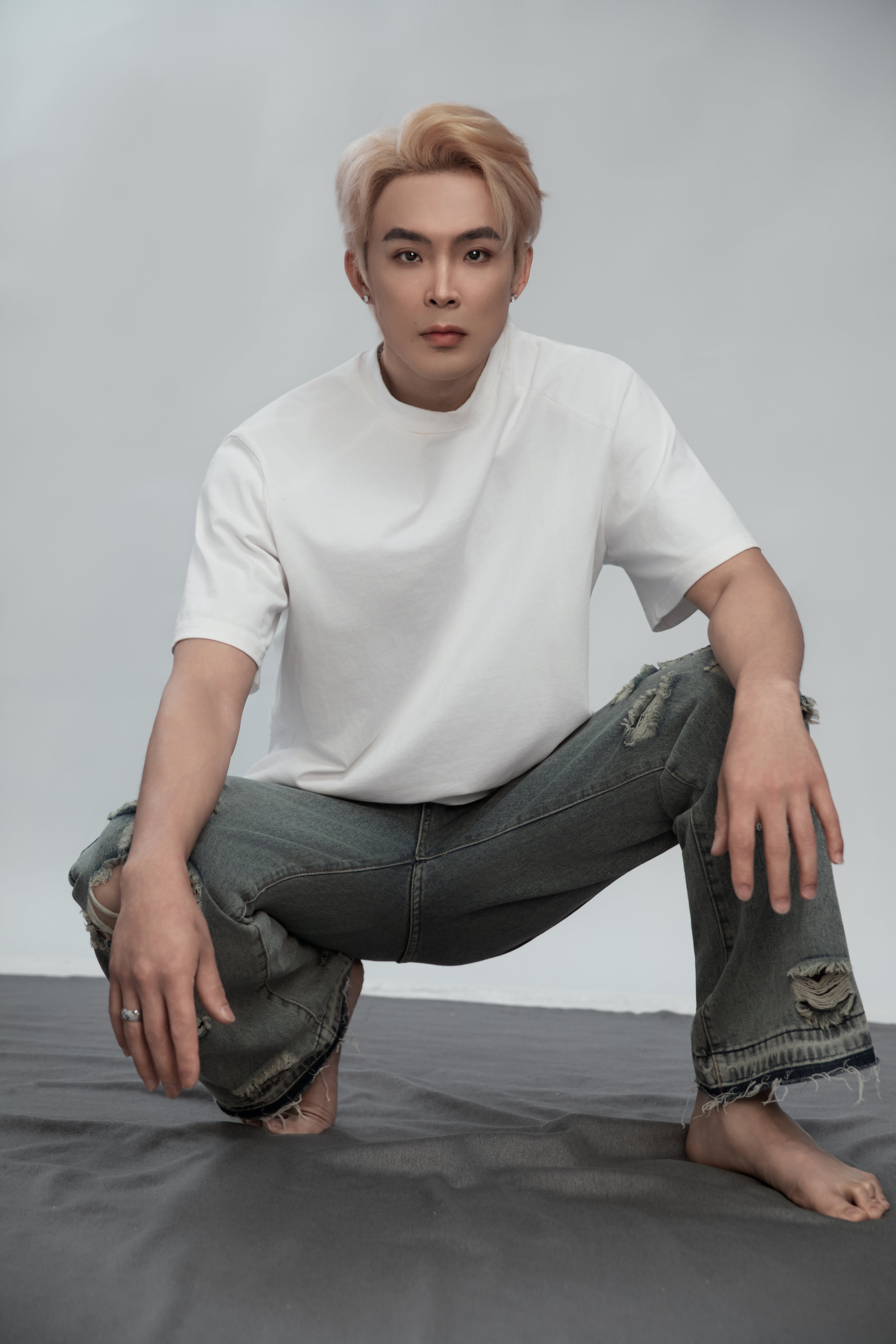  A man in a white shirt and jeans crouches confidently, posing for the camera against a white backdrop with a gray rug beneath him.