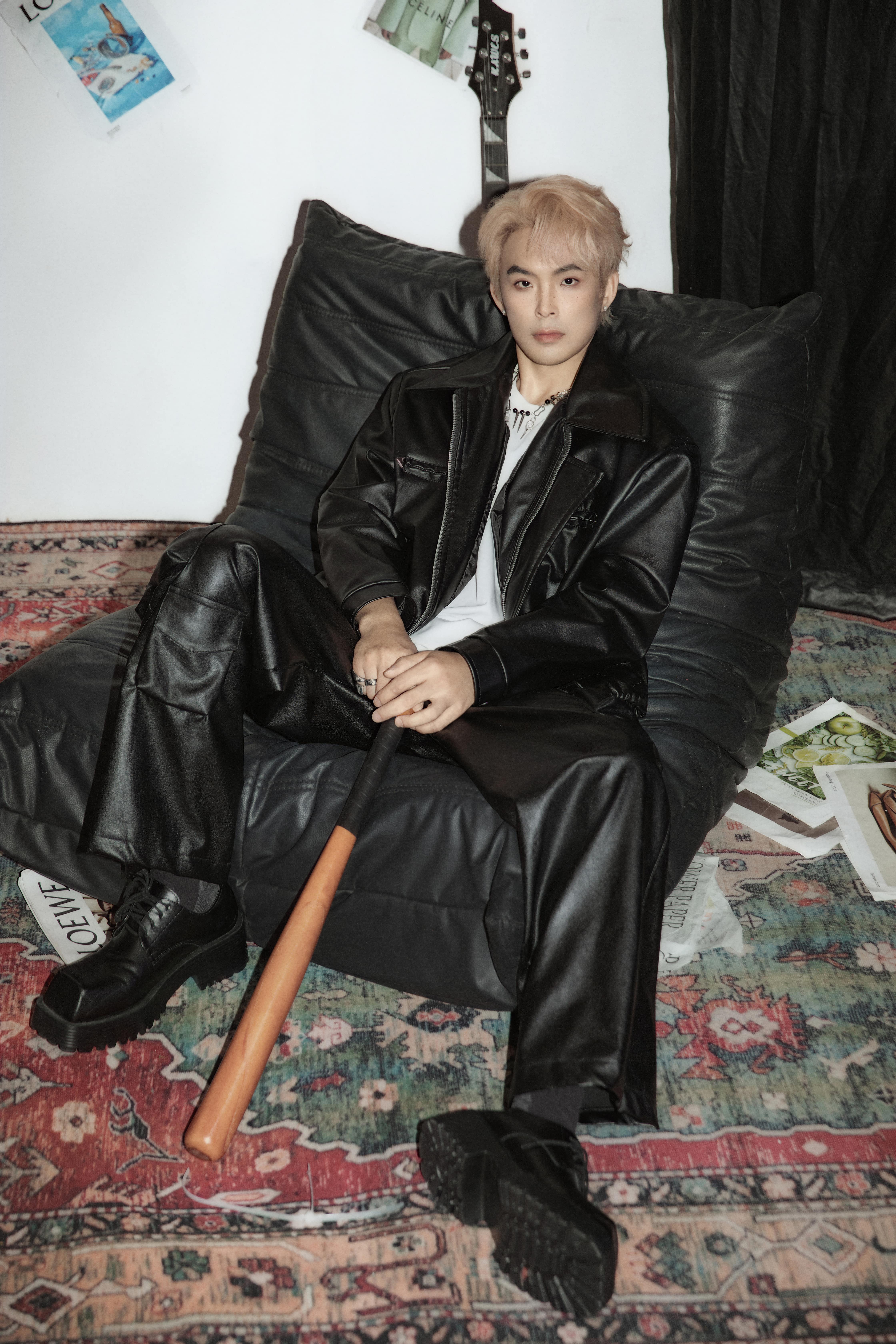 A man rests on a black fuzzy chair, looking up at the camera in front of a white background with magazines and posters.