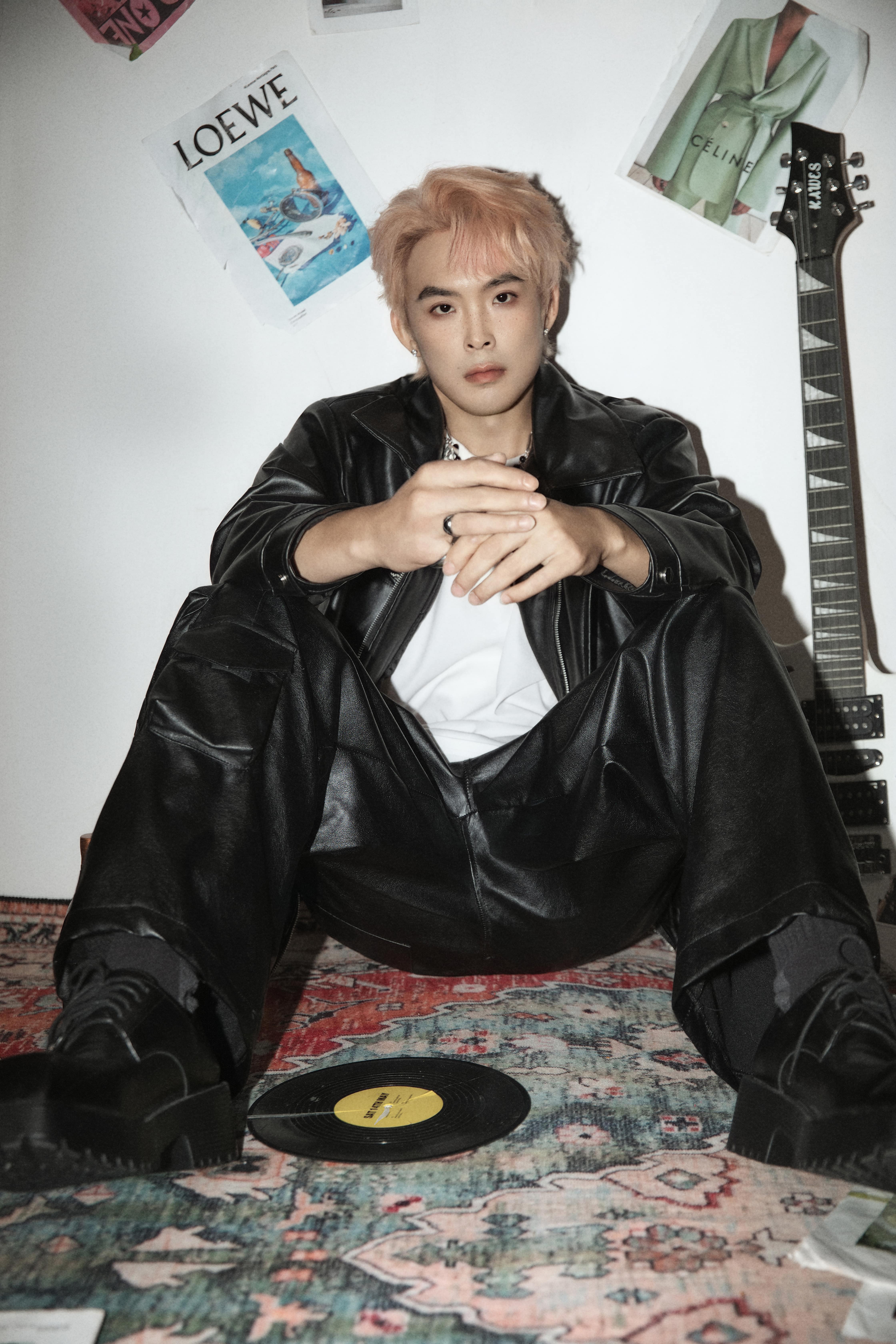 A man sits facing the camera, holding his hands together, in front of a white background with magazines and posters.