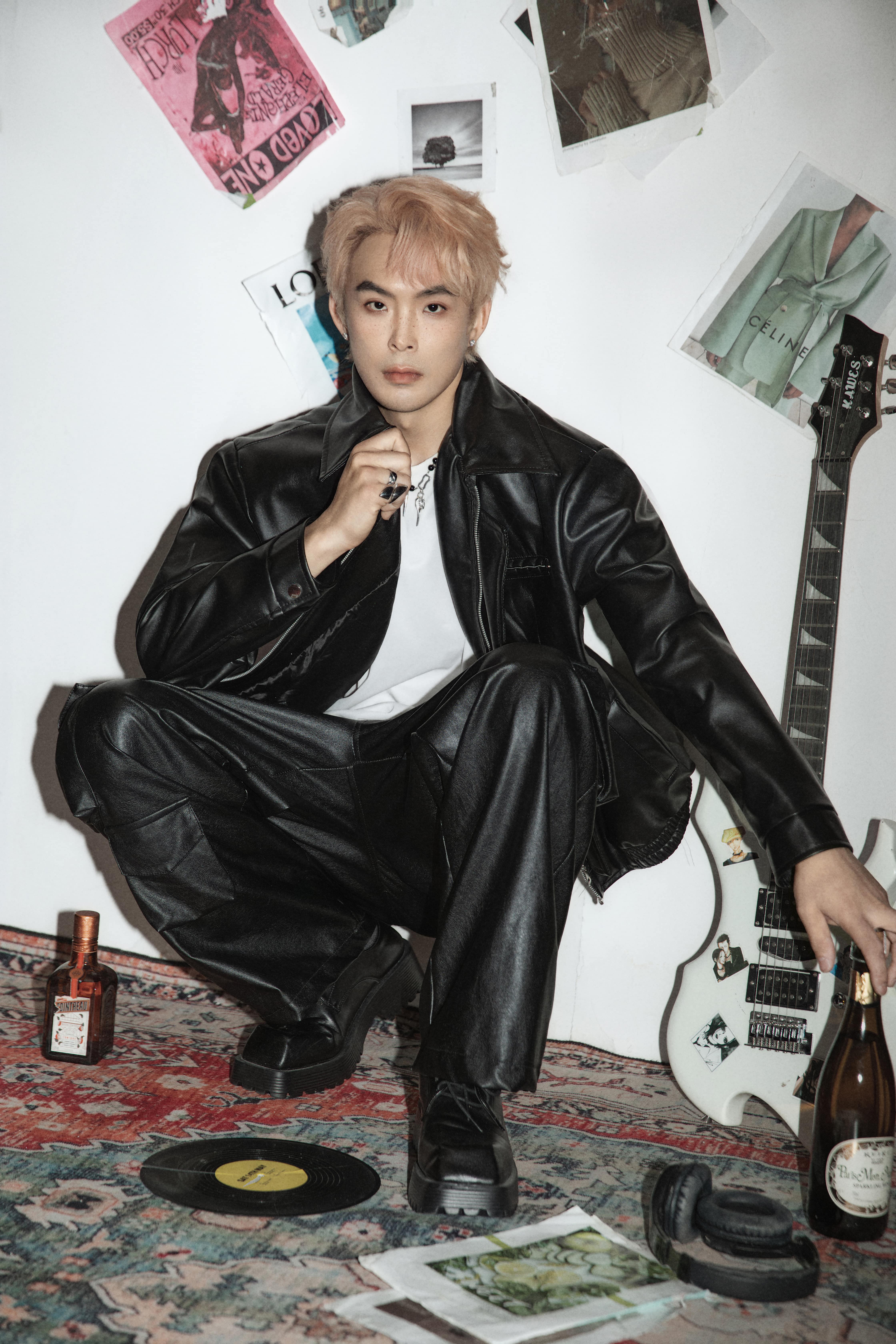 A man crouches, resting his hand on an alcohol bottle, in front of a white background with magazines and posters.