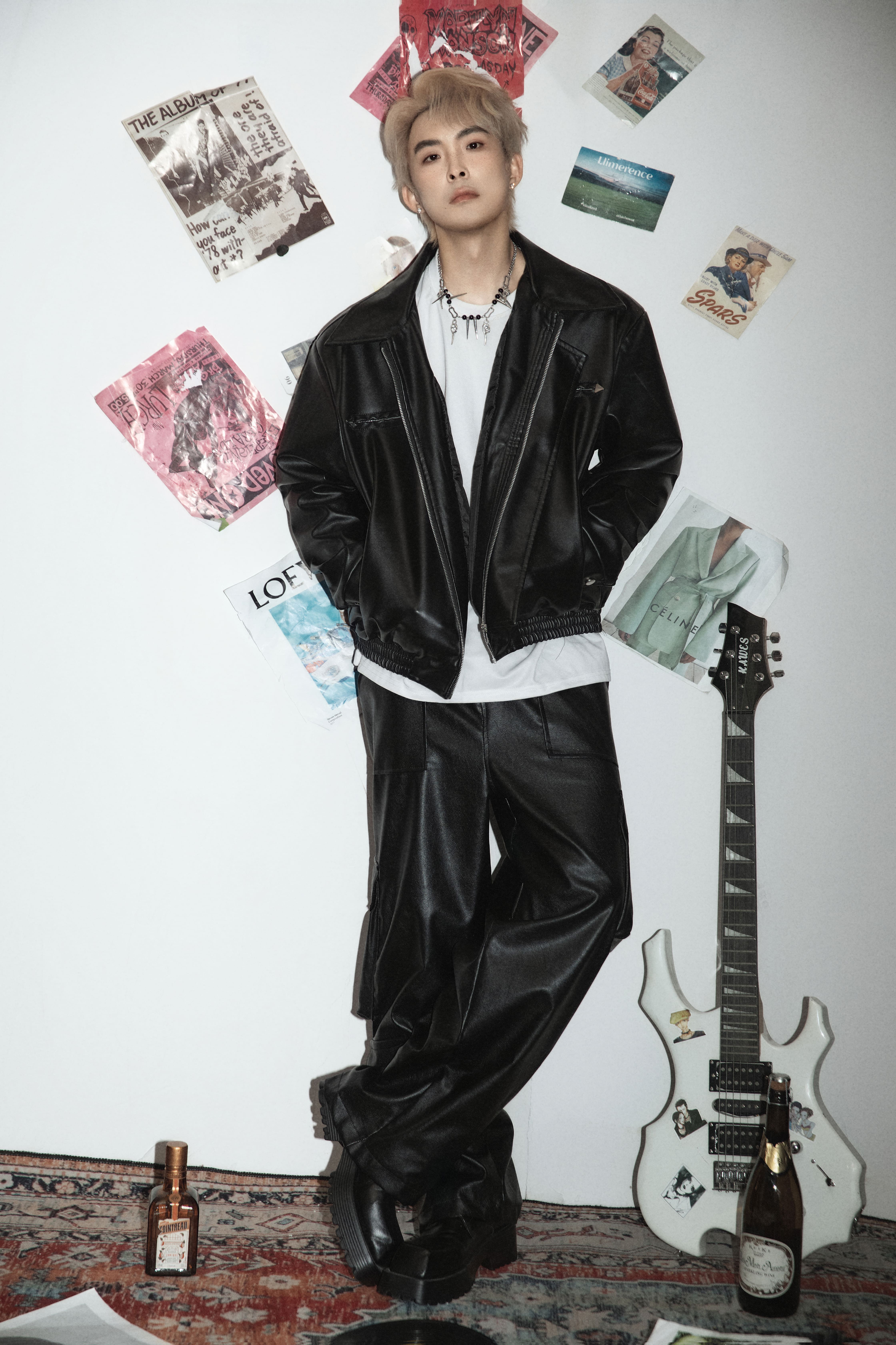 A man stands with one leg crossed over the other against a white background with magazines, posters, a guitar, alcohol bottles, and a record on the floor.