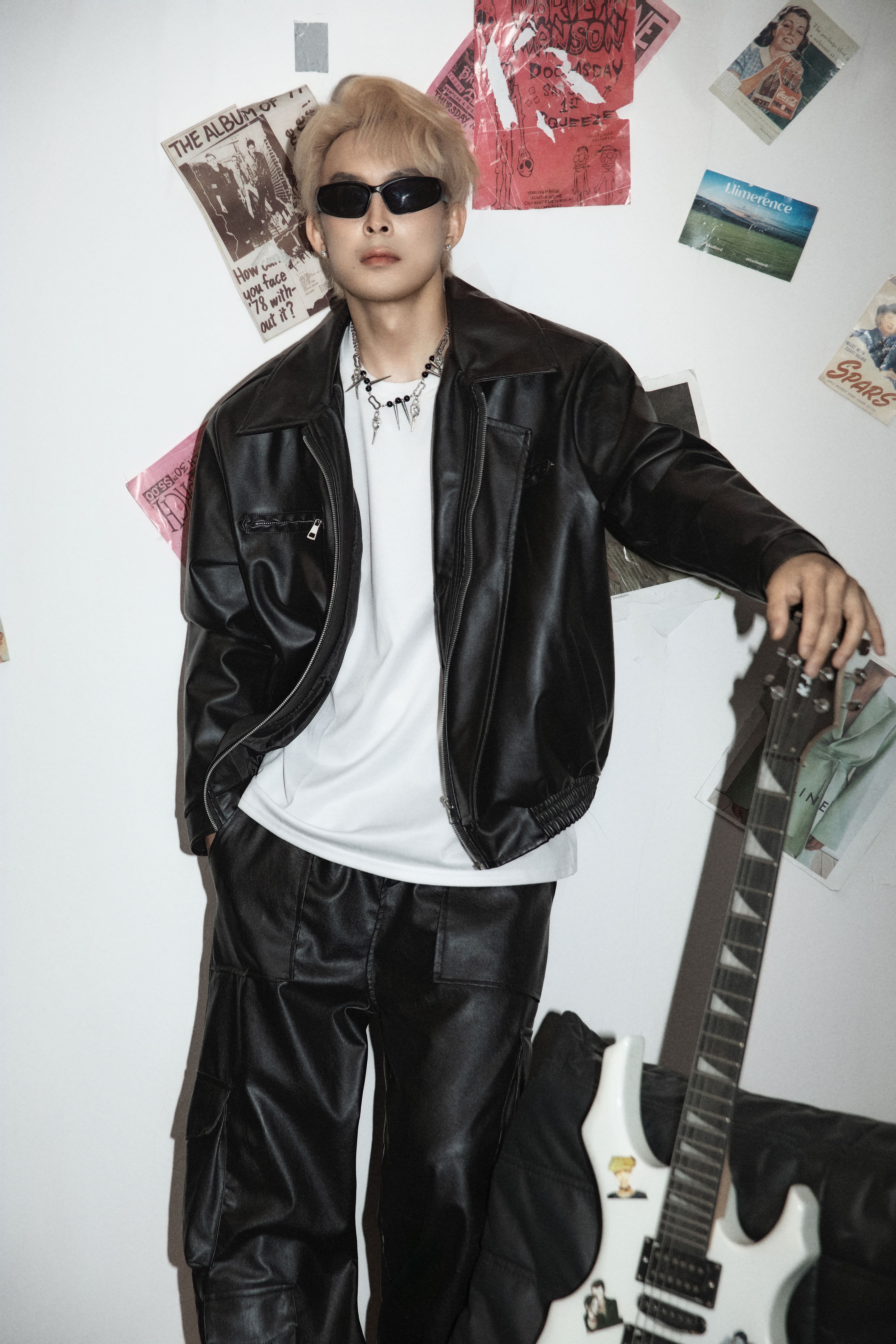 A man poses with his arm resting on a guitar in front of a white background with magazines and posters.