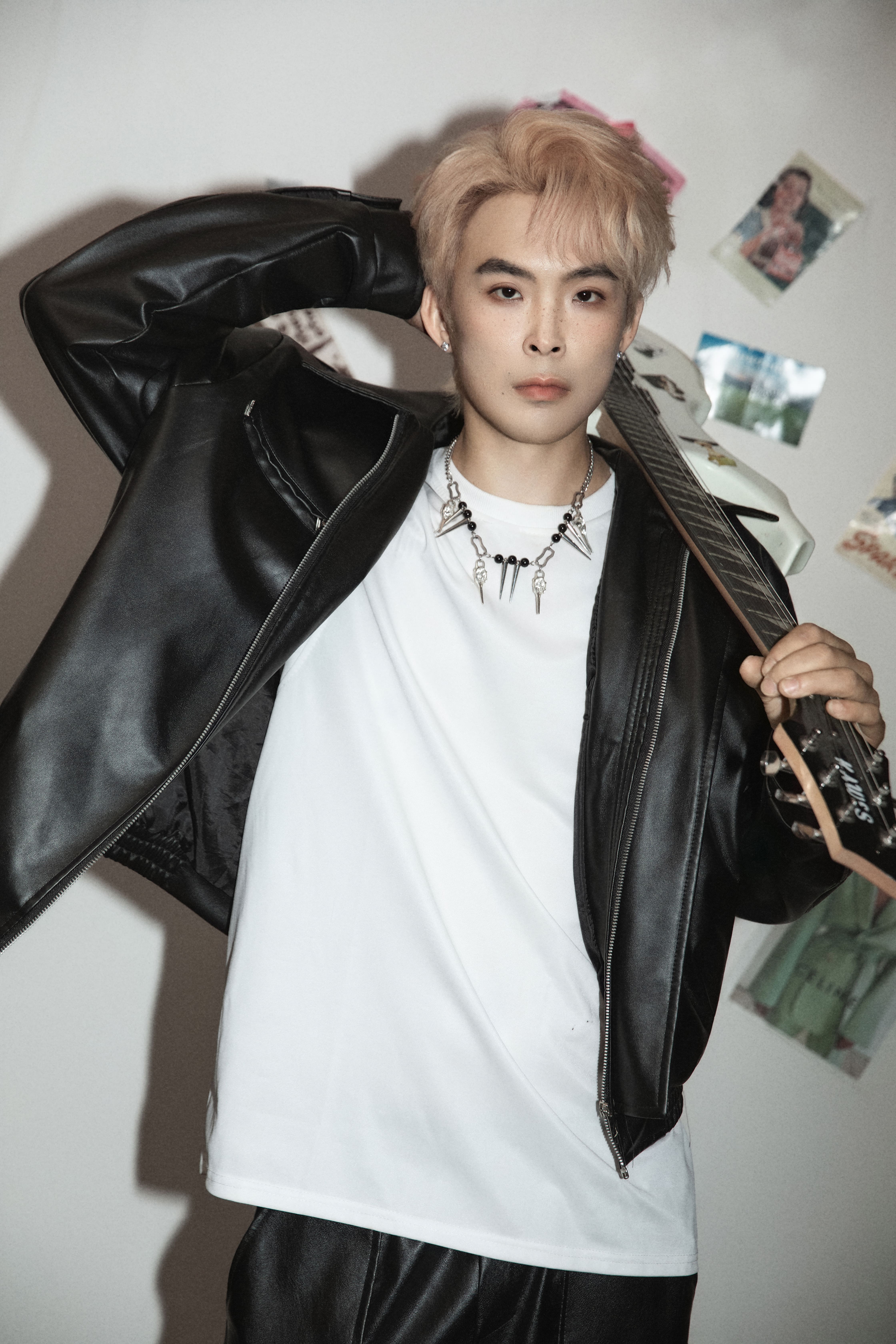 A man poses in front of a white background with magazines and posters, holding his neck and a guitar on his shoulder.