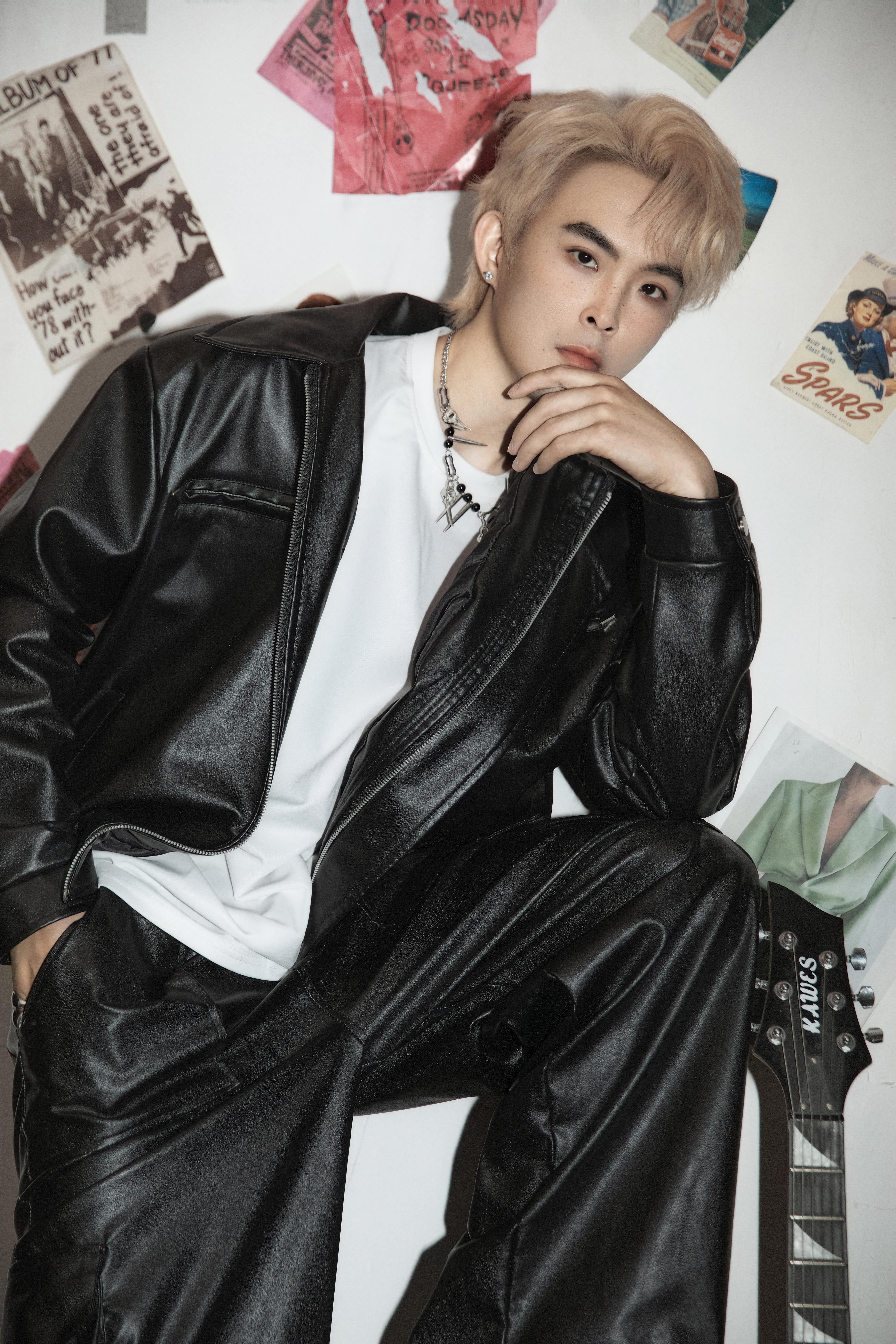 A man rests with one leg elevated, one arm on his knee, and his head supported by his hand against a white background with magazines and posters.