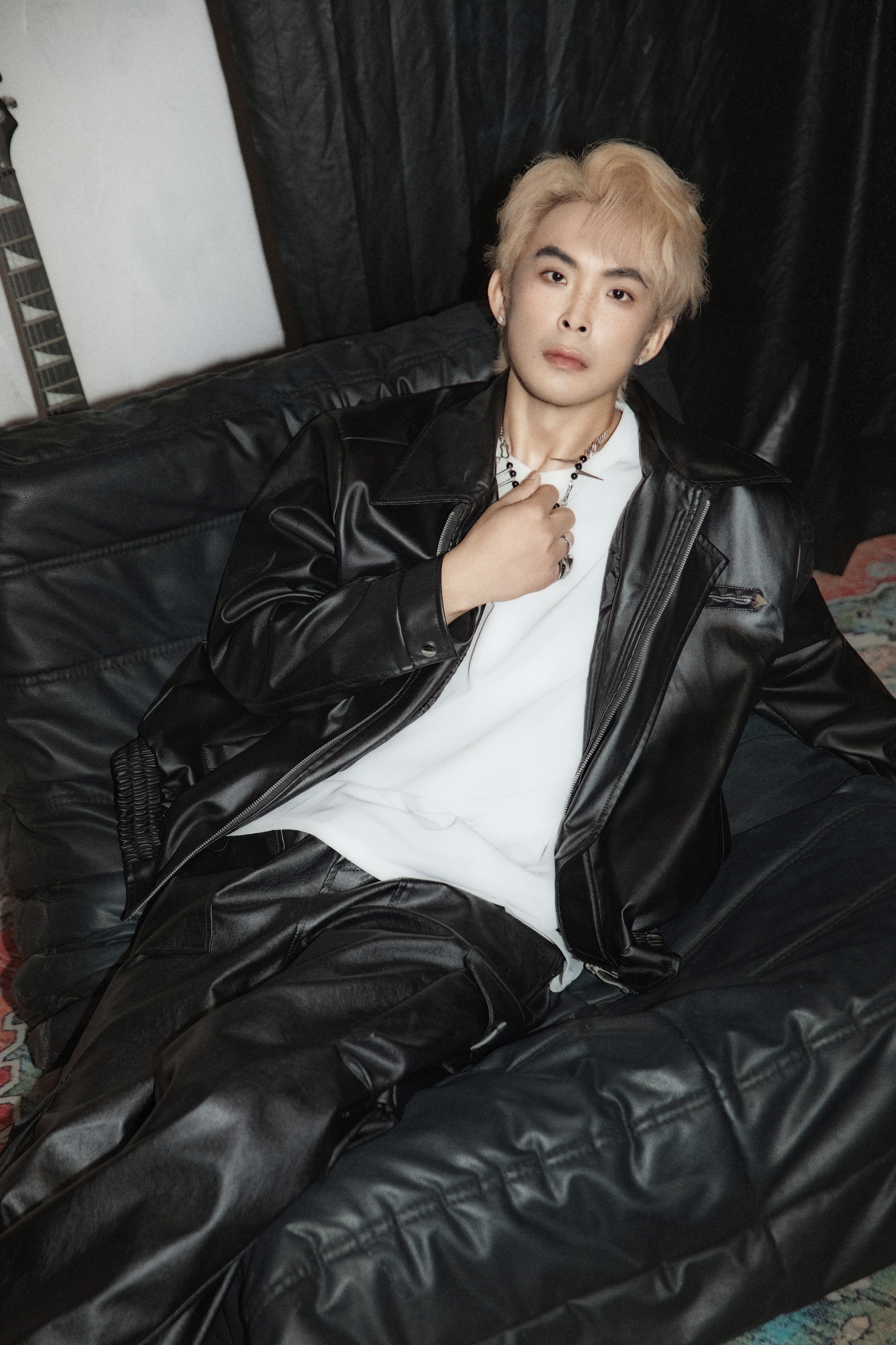 A man rests on a black fuzzy chair, playing with a chain in front of a white background with magazines and posters.