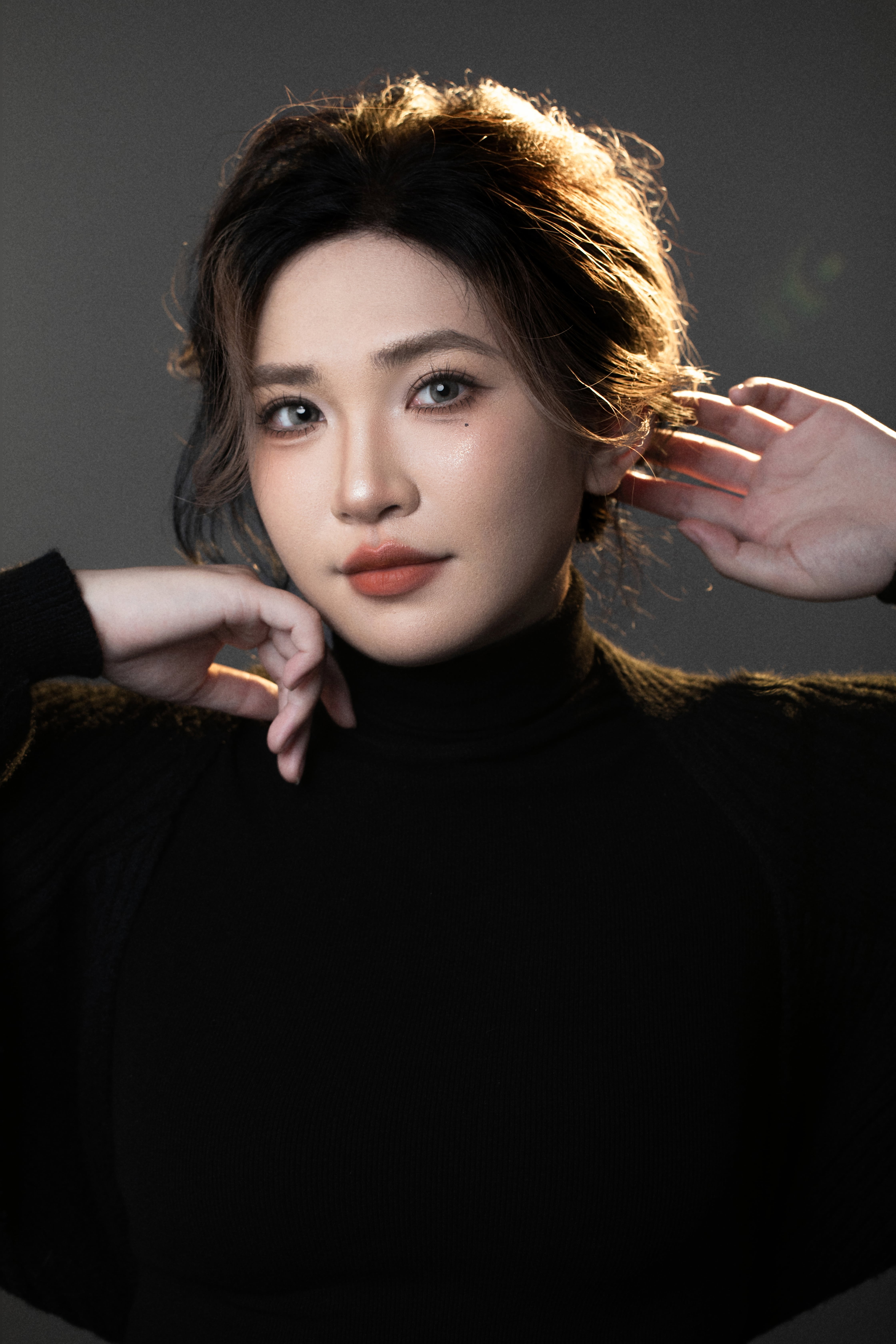 A woman rests one hand behind her head and the other near her chin in a close-up.”