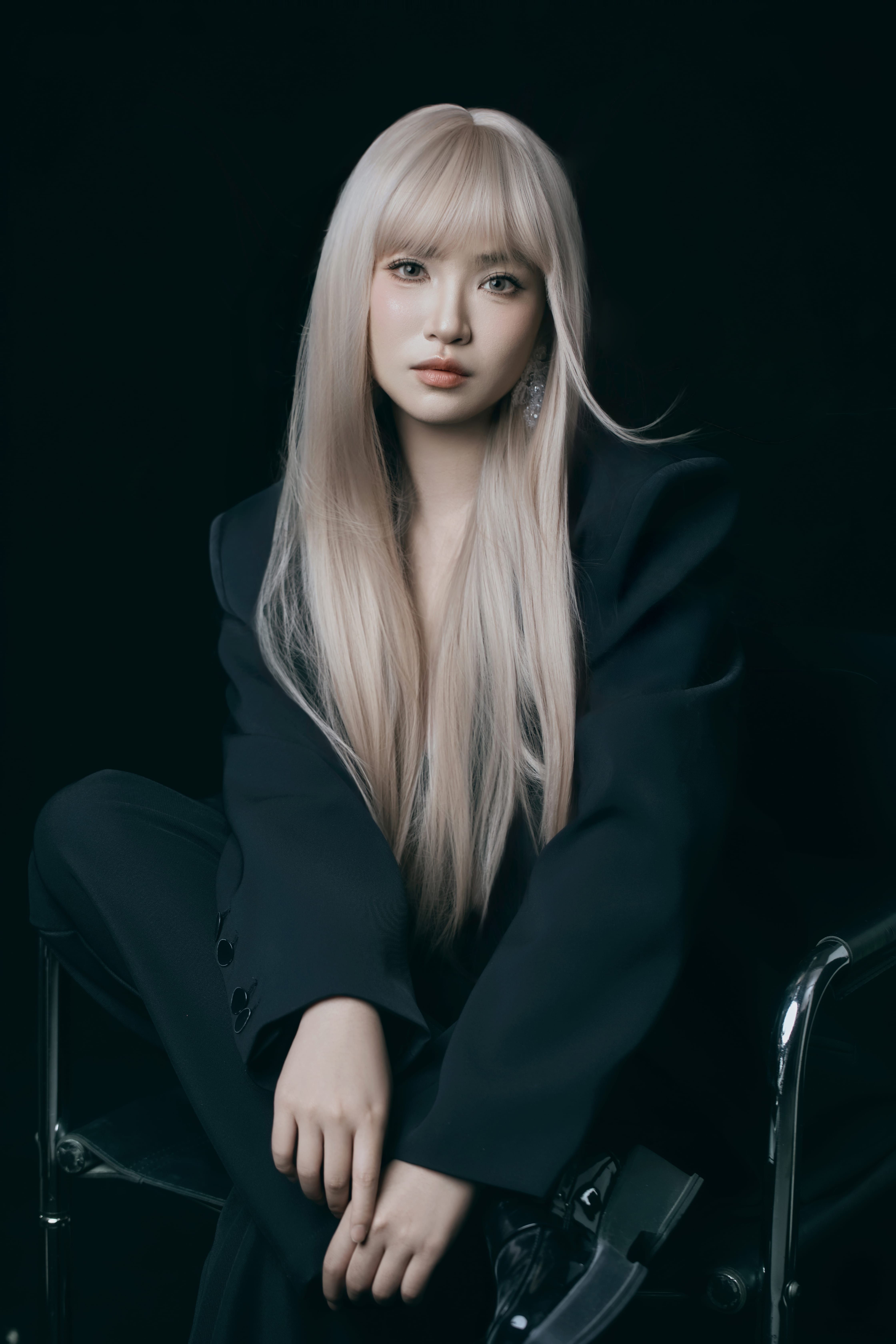 A woman poses seated on a chair, one leg crossed over the other, facing the camera.