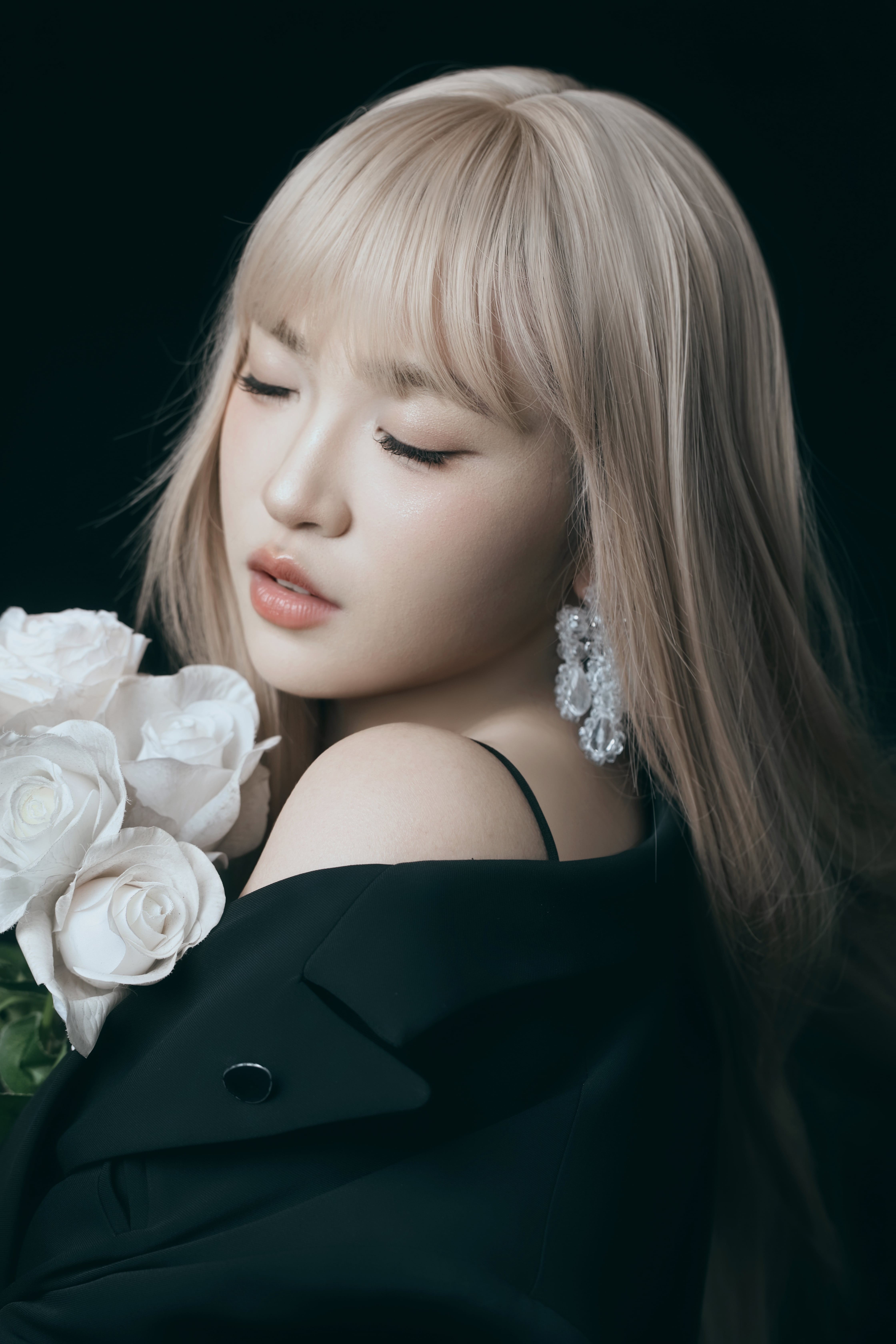 A close-up of a woman with eyes closed, holding flowers in front of a black background, with wind sweeping her hair.