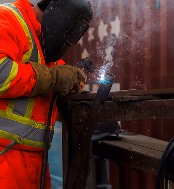 Worker in a high-visibility orange safety suit and welding helmet welding a metal pipe with sparks and smoke.