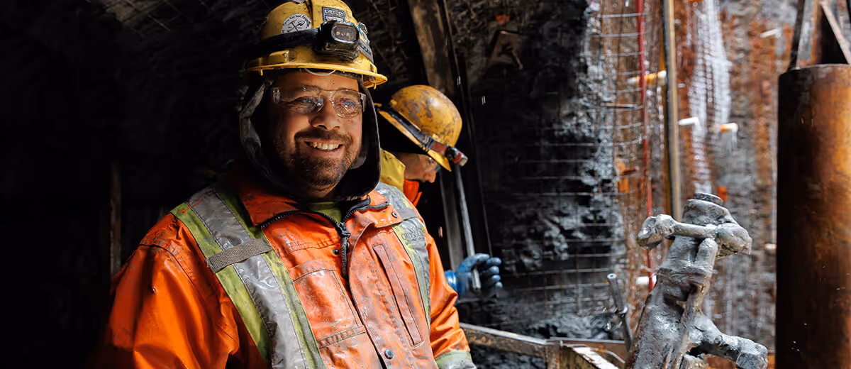 Smiling miner in orange safety jacket and yellow helmet inside a dark tunnel with another miner working behind him.