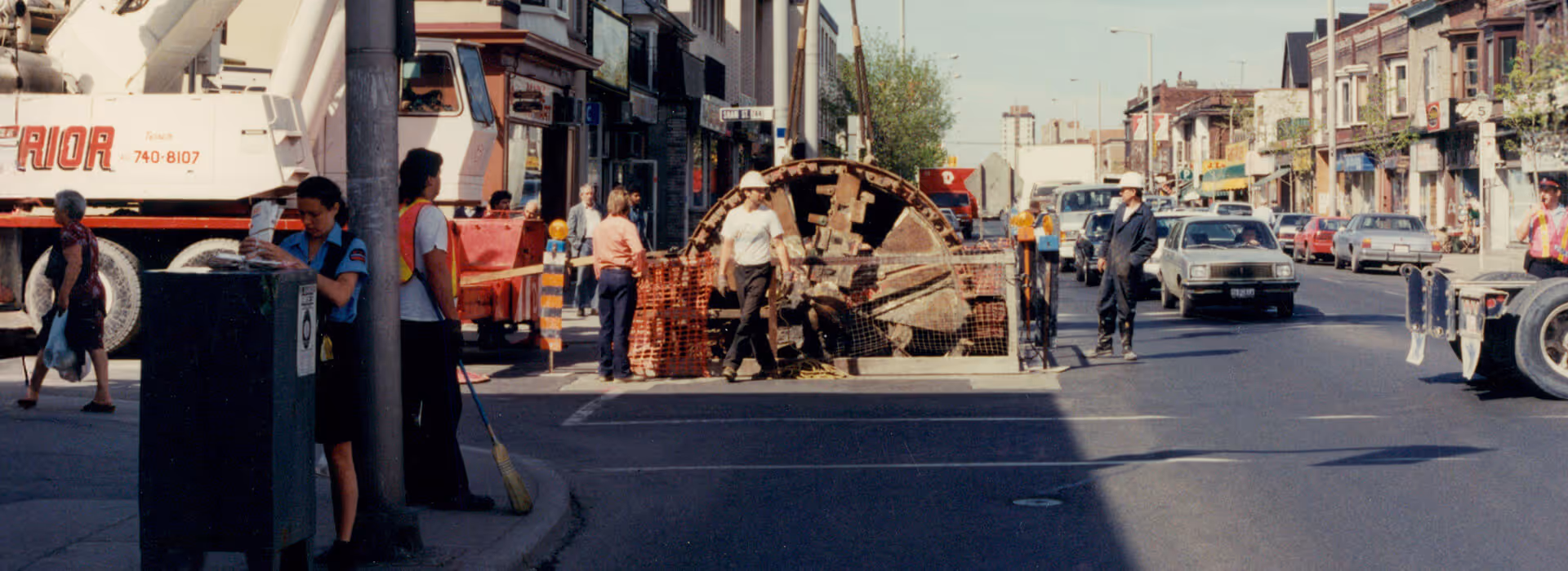 Street scene with construction workers near a large tunnel boring machine on a city road, accompanied by a police officer and passing cars.