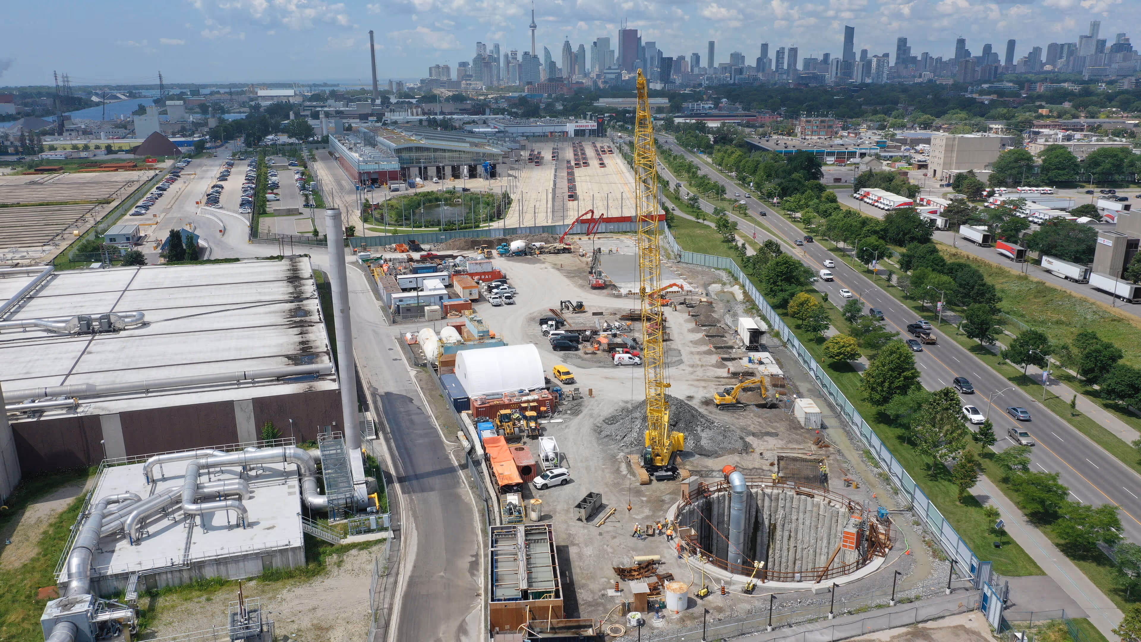 Aerial view of a construction site with a yellow crane and deep excavation, set against a city skyline with tall buildings and a cloudy sky.