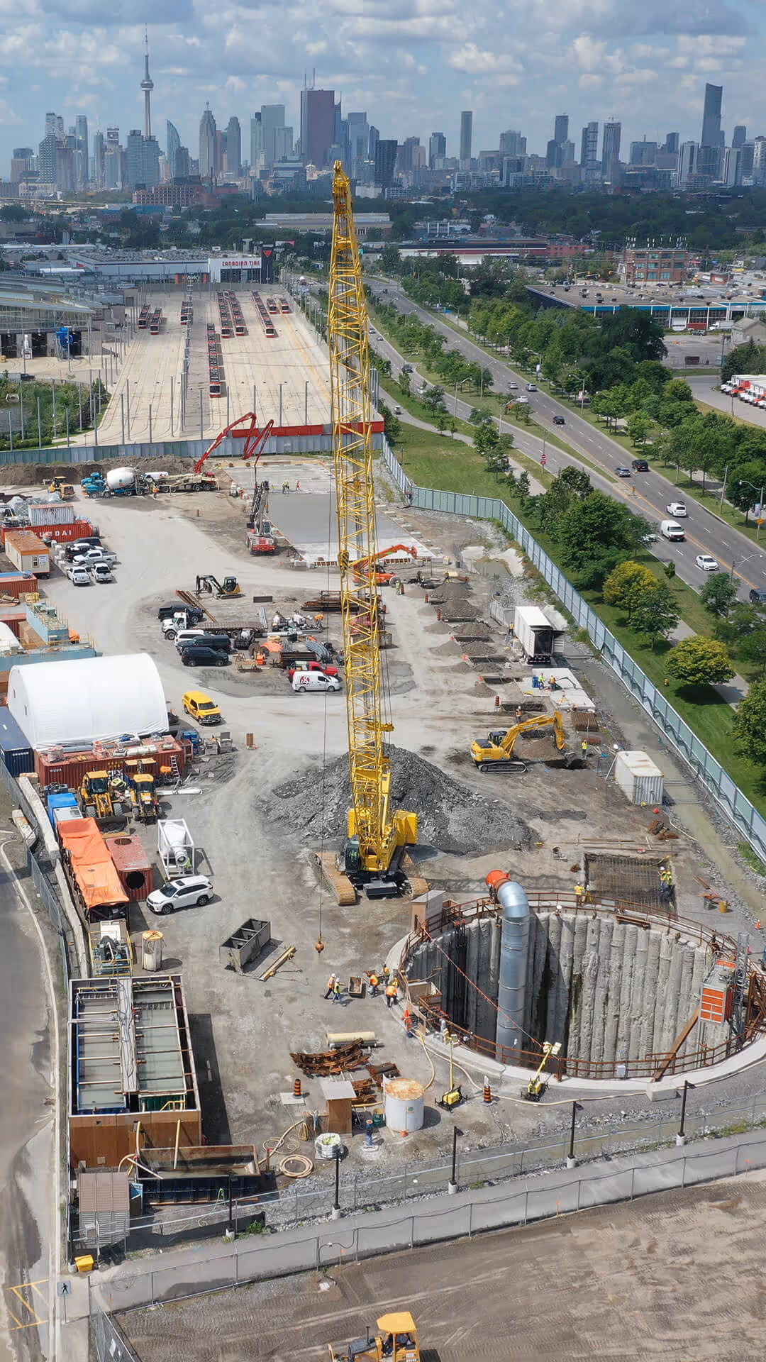 Construction site with a large yellow crane, workers, machinery, and a deep circular excavation, with a city skyline in the background under a cloudy sky.