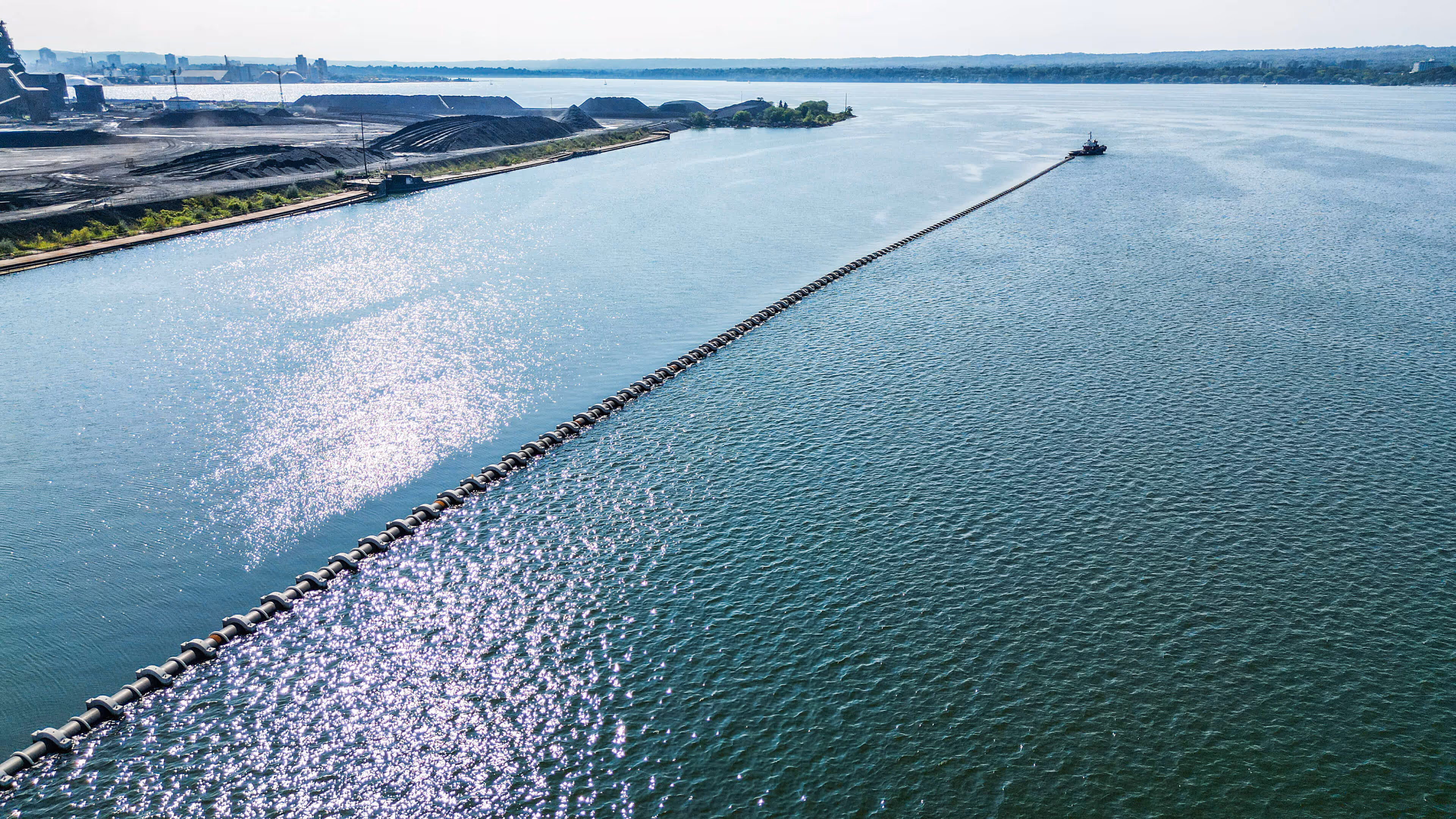 Large body of water separated by a long floating barrier with industrial buildings and piles of coal on the left shore under a clear sky.
