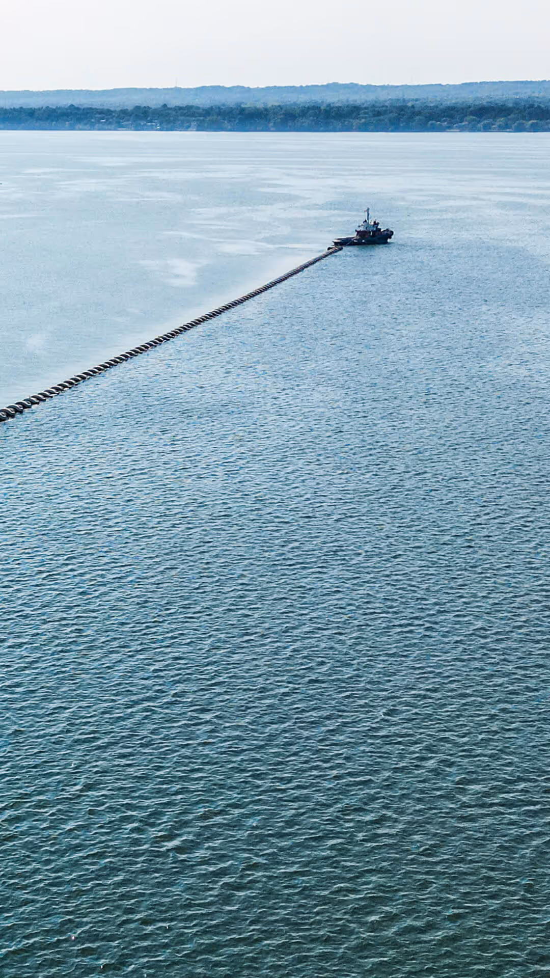 Tugboat pulling a long line of floating buoys across a large body of water with a distant forested shoreline.
