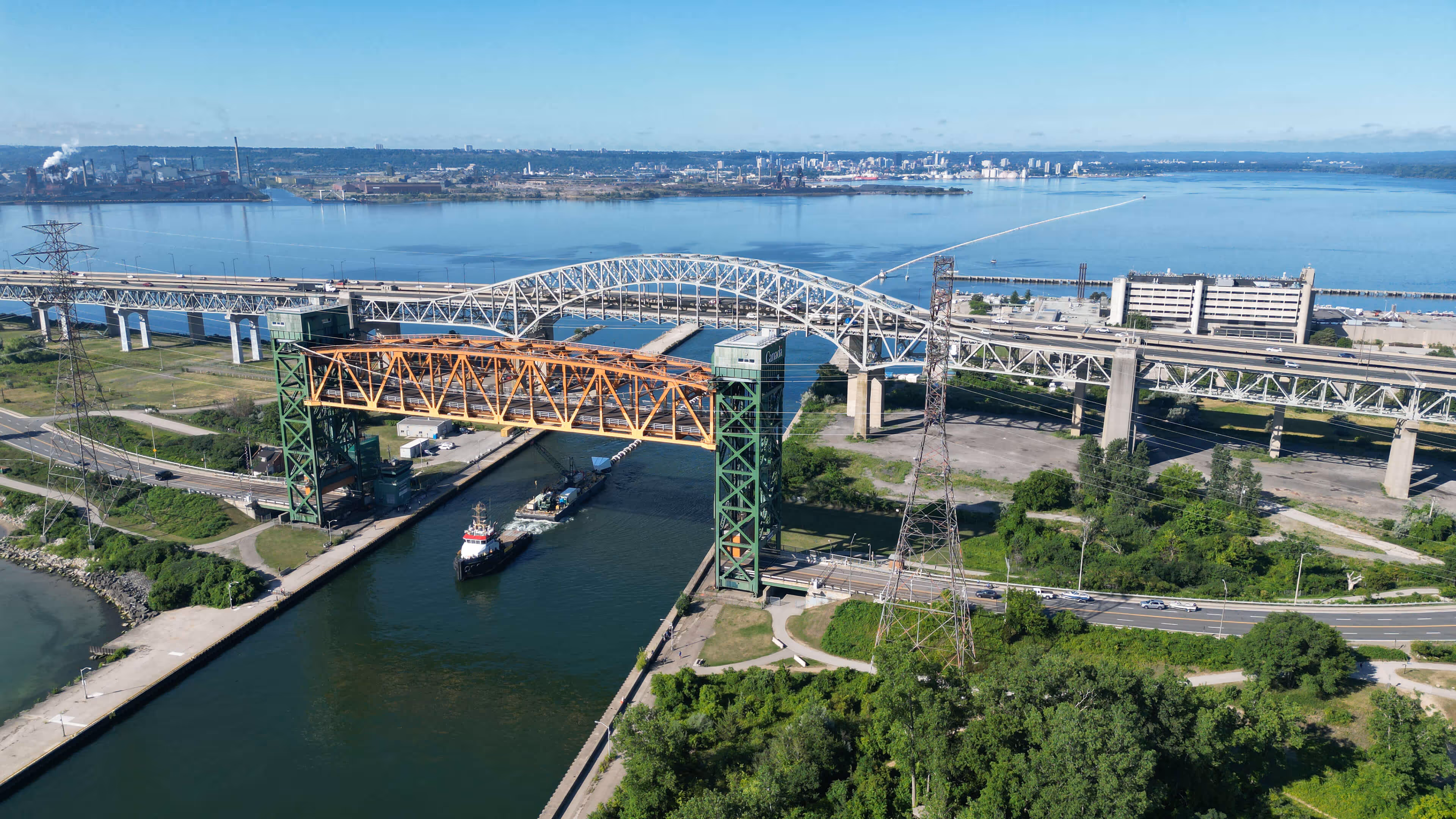 Aerial view of two bridges crossing a waterway with a tugboat passing underneath, surrounded by greenery and urban buildings in the background.