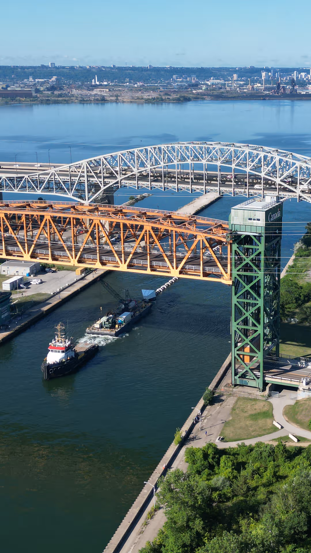 A green lift bridge with an orange truss section over a river, with a ship passing underneath and a city skyline in the background.