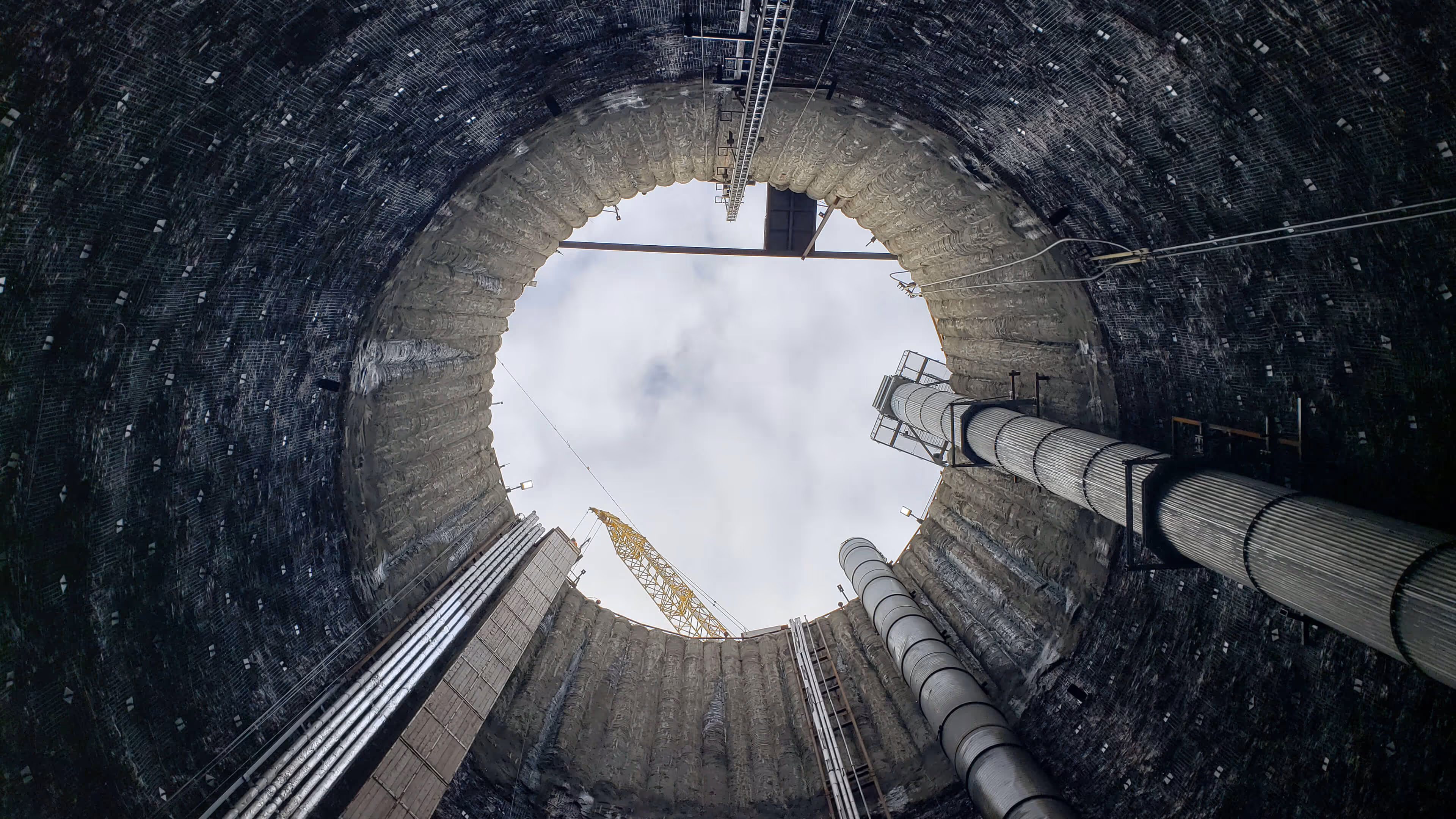 View looking up from inside a large industrial cylindrical structure with pipes, scaffolding, and a crane visible against a cloudy sky.
