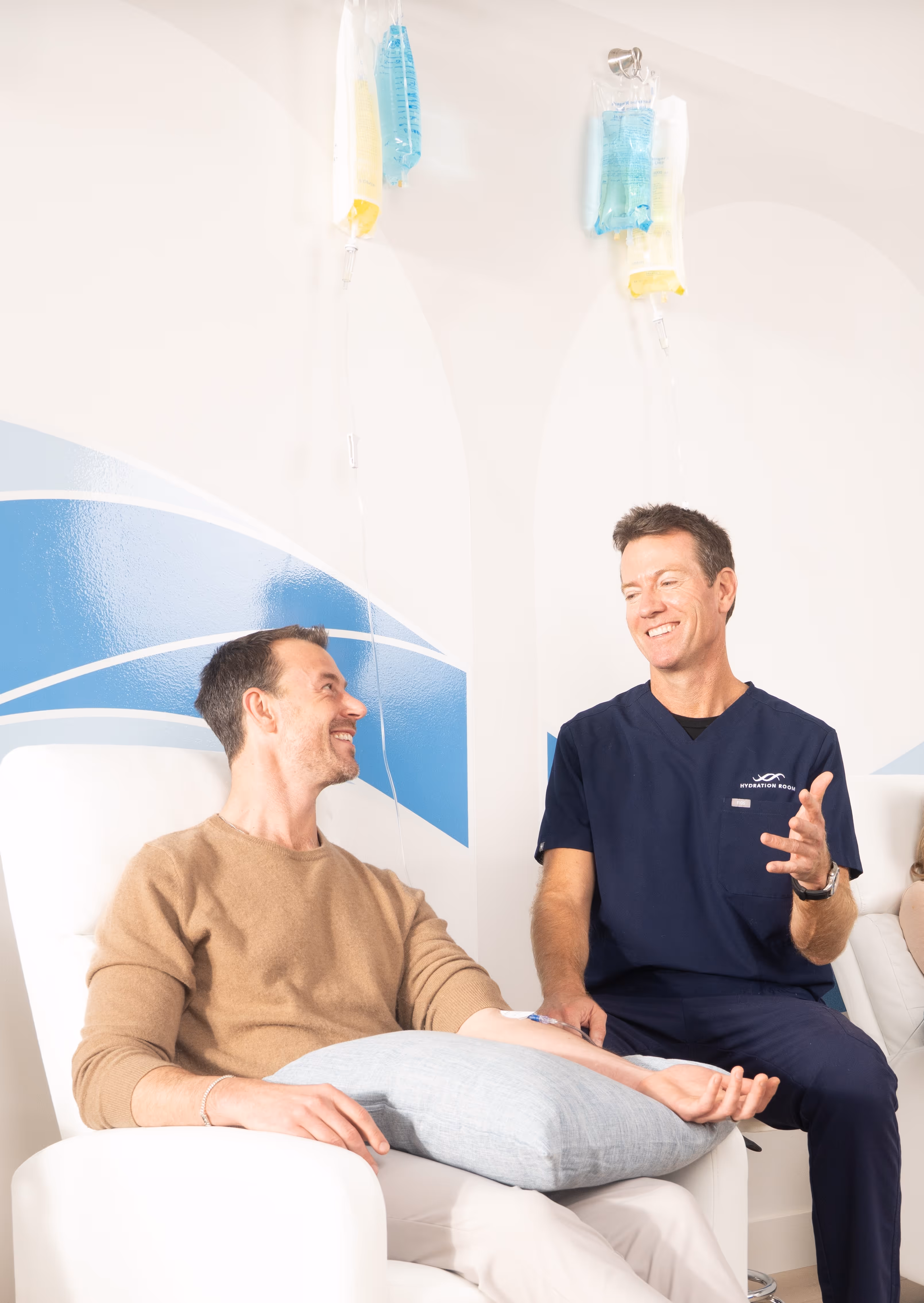Medical professional in navy scrubs talking and smiling with a seated male patient receiving an IV infusion in a bright room.
