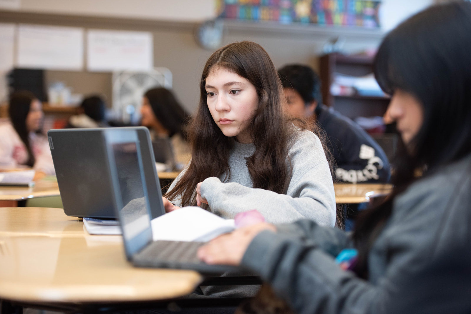 Teenage students in a classroom working on laptops, focused on their screens with blurred students in the background.