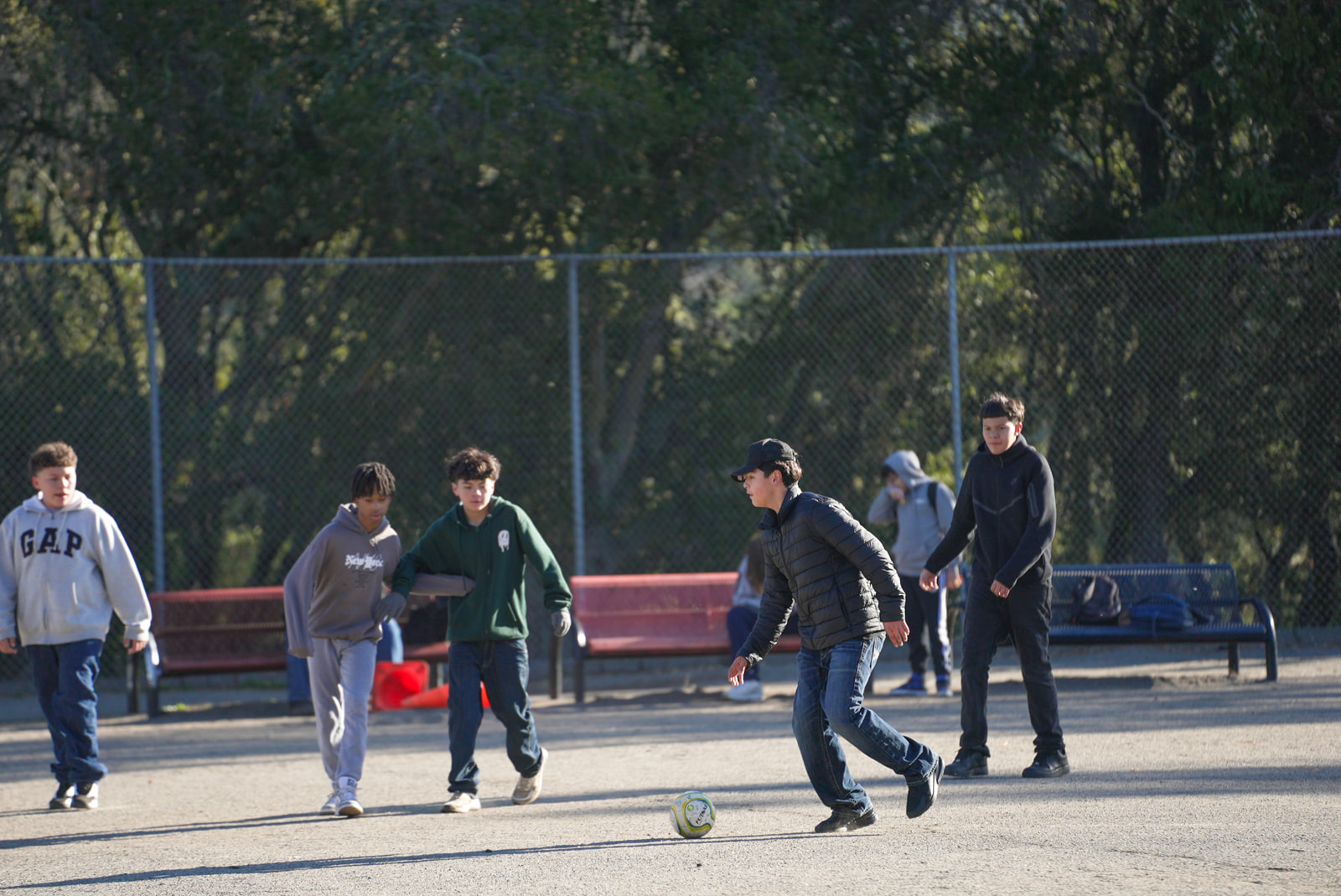 Five boys playing soccer on a dirt field with a chain-link fence and trees in the background.