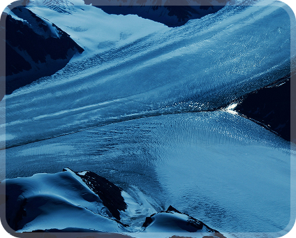 Aerial view of glacier flowing between snow-covered mountain peaks under blue light.