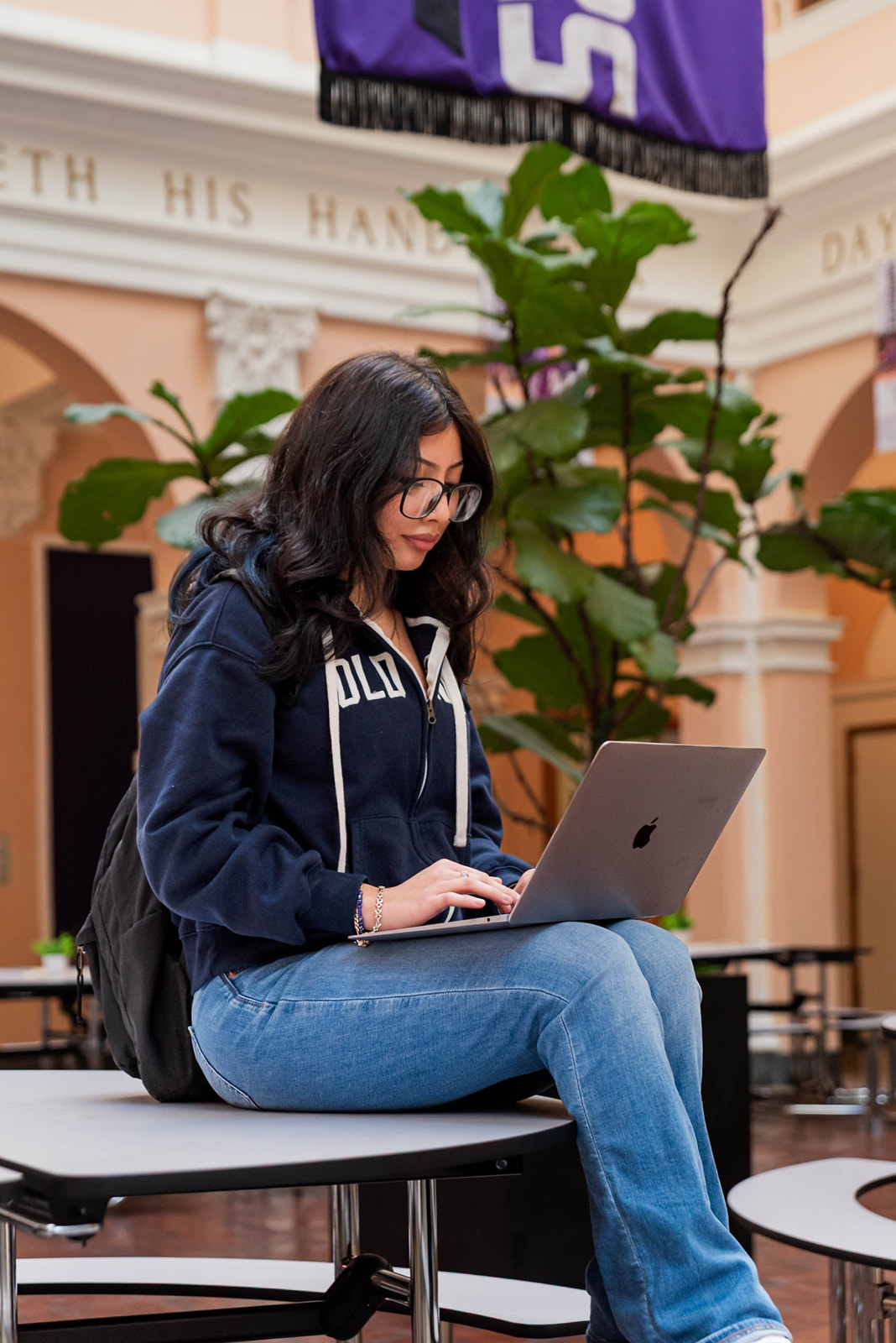A smiling young person with short curly hair sits at a desk, holding a pen and wearing a blue Adidas hoodie with headphones around their neck.